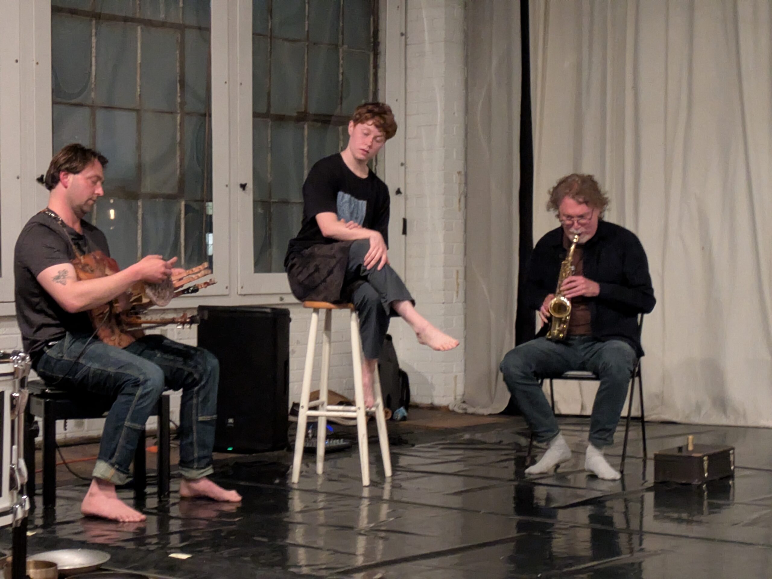 Three people sit in an oblique triangle that fills the frame. To the left, a musician, Aabeizer, sits on a black bench in carpenter jeans and a dark t-shirt. His eyes are closed and his feet bare. He moves his hands around a circular plate and wooden dowels that extend from a wood board he holds against his chest. To the right, a saxophonist, Bhob Rainey, sits on a folding black chair in a black cardigan and grey pants, blowing into the mouthpiece and pressing the keys. Between them, a person with short red curls, Kayliani Sood, crosses their legs on a white stool, sitting higher than the musicians beside her. They wear brown shorts over grey pants and a black t-shirt with a blue square patch in the center. She rests one hand on her knee, and the other over their forearm, closes her eyes and tilts their head pensively to the right.