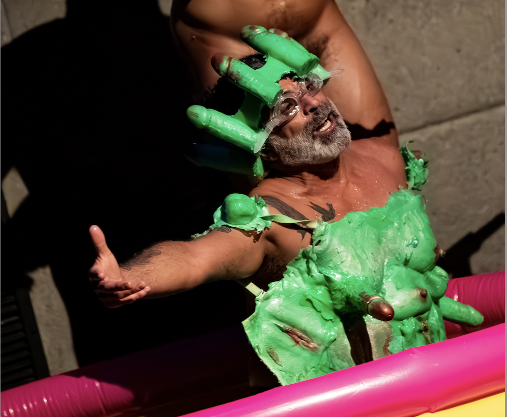 Antonio Ramos, a brown man with a full, greying beard sits in a kiddie pool under Saúl Ulerio, another brown man with a greying head of hair. They both are naked, Ulerio pumping baby oil down onto Ramos from a squirt bottle, who wears goggles, a poisonous, garish green top, and a headband, all made of penis and breast molds. Ramos extends an arm out to the left side of the image while speaking to the audience.