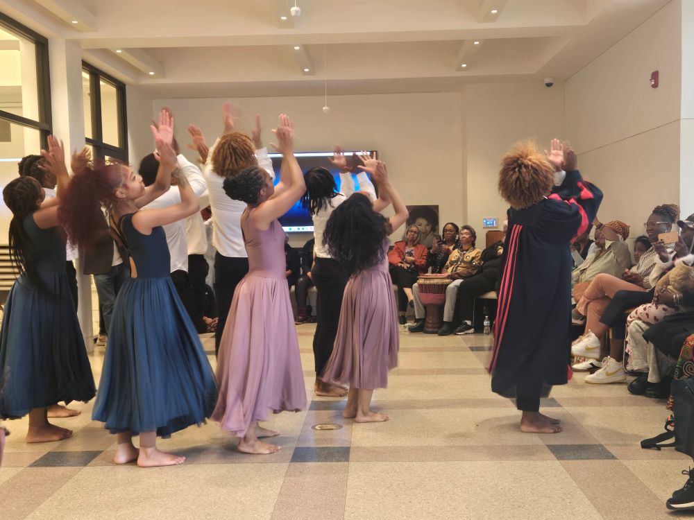 The West Philadelphia High Dance Ensemble performs in front of an audience seated around the perimeter of the room. The dancers stand close to each other with their arms raised mid-clap overhead. Some dancers wear long evergreen or rose colored dresses, while others are dressed in black pants and a white button-down shirt. One dancer stands in front of the group wearing a preacher’s robe. The ensemble resembles a lively church congregation.