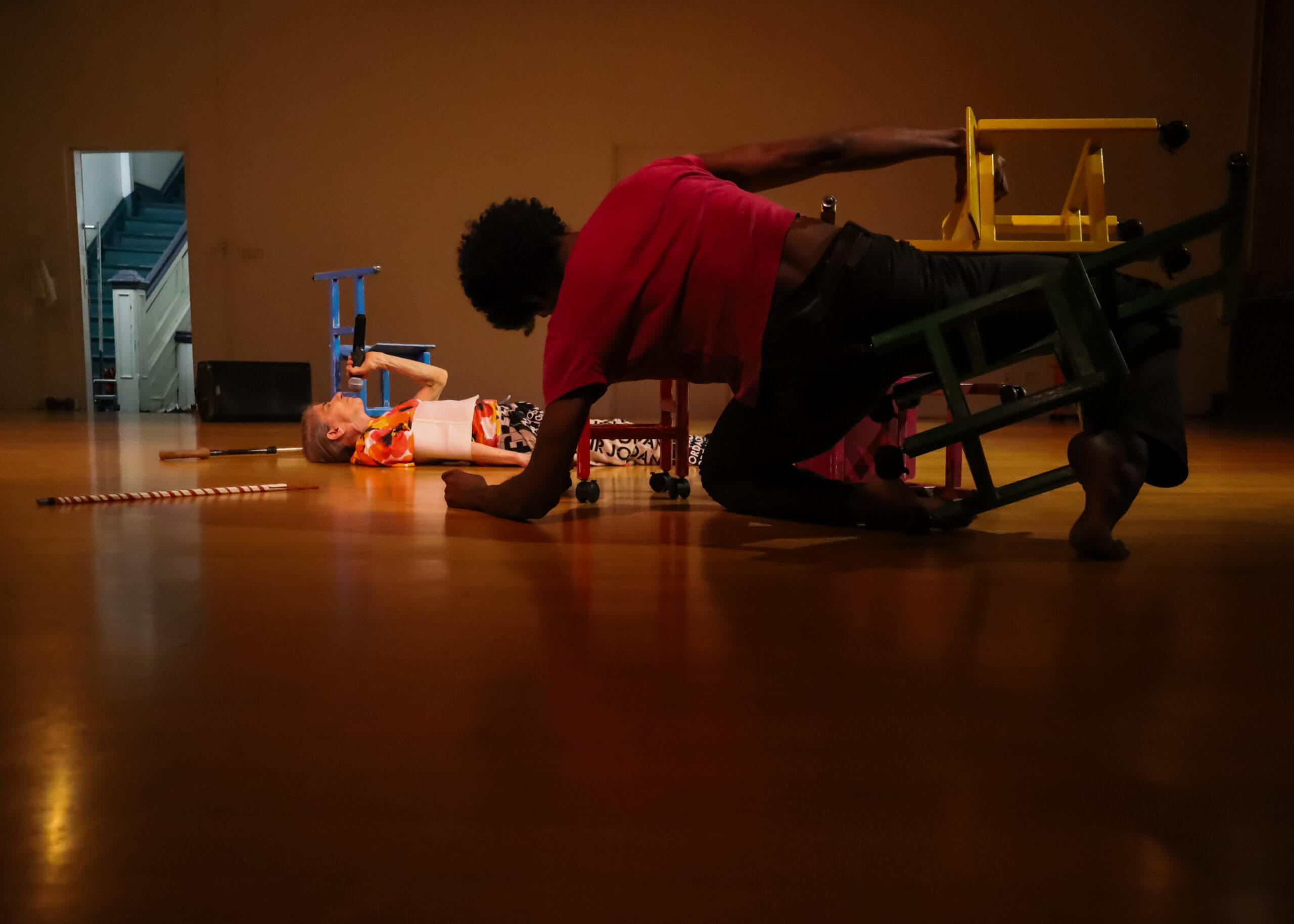 In the foreground and in darkness, dancer Zo Williams is balanced on his left forearm and left shin. In between his legs is a tiny wooden chair, beneath. another yellow one he holds in place with his right hand. Zo faces upstage, where Cathy Weis lies on the floor on her back, with what looks like two canes lying on the floor on either side of her head. More brightly illuminated, Cathy holds a microphone up to her mouth, a tiny blue chair beyond her. In the distance, through an open door to the hallway, we see stairs going up.