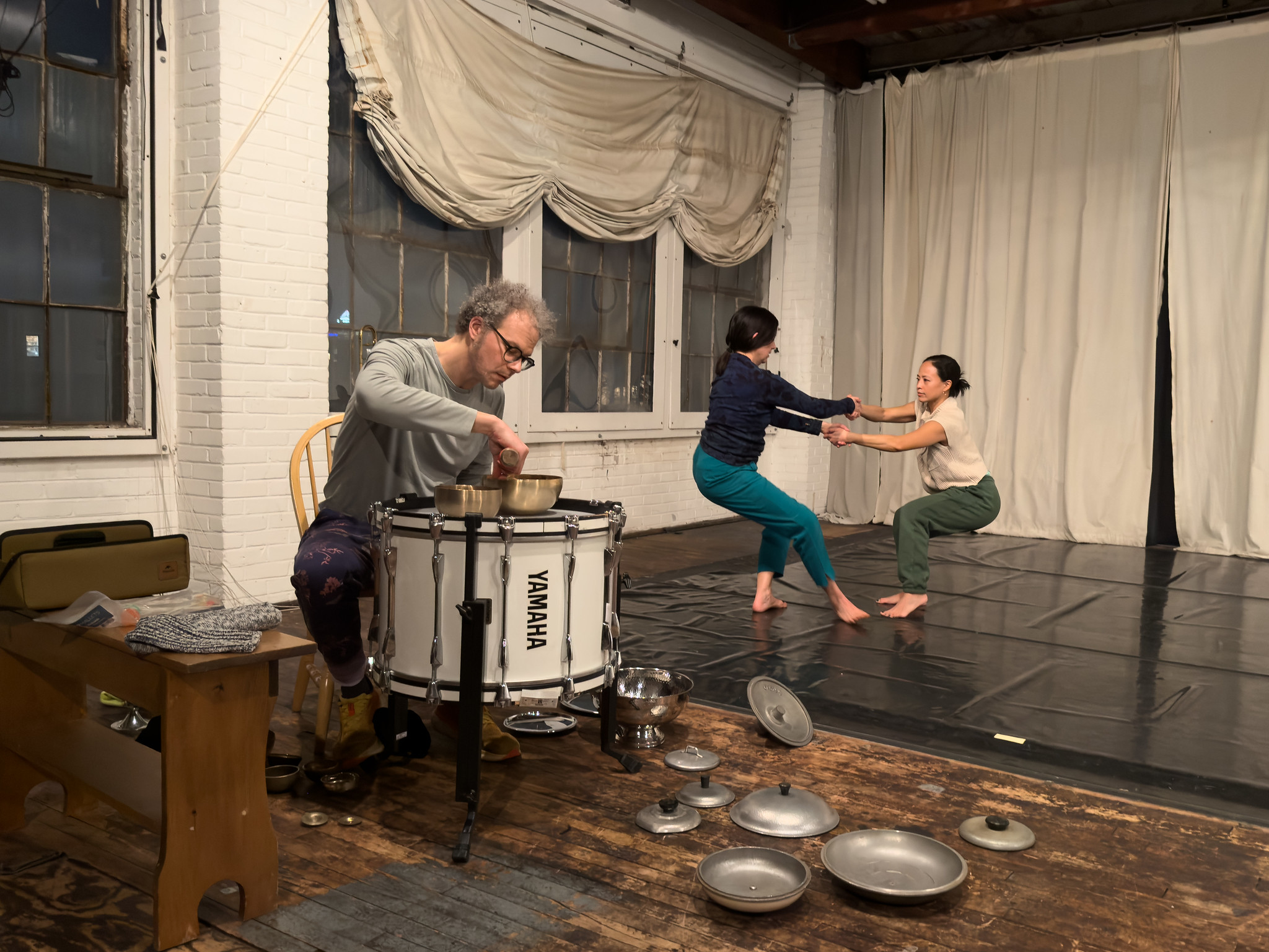 In the foreground, a percussionist, Flandrew Fleisenberg, wearing a grey long sleeve and glasses leans over and rings a brass soundbowl that he’s placed on top of a big white YAMAHA drum. Metal kitchen pot lids are strewn on the faded wood floor around him. In the background, a person in teal pants and a navy longsleeve, Loren Groenendaal, crosses arms and holds hands with a person wearing green pants and an off white t-shirt, Michele Tantoco. They counterbalance, sending their hips back and holding each other's gaze. Beneath their blurring feat, black marley crinkles, behind and beside them, white curtains drape the backstage and the windows.