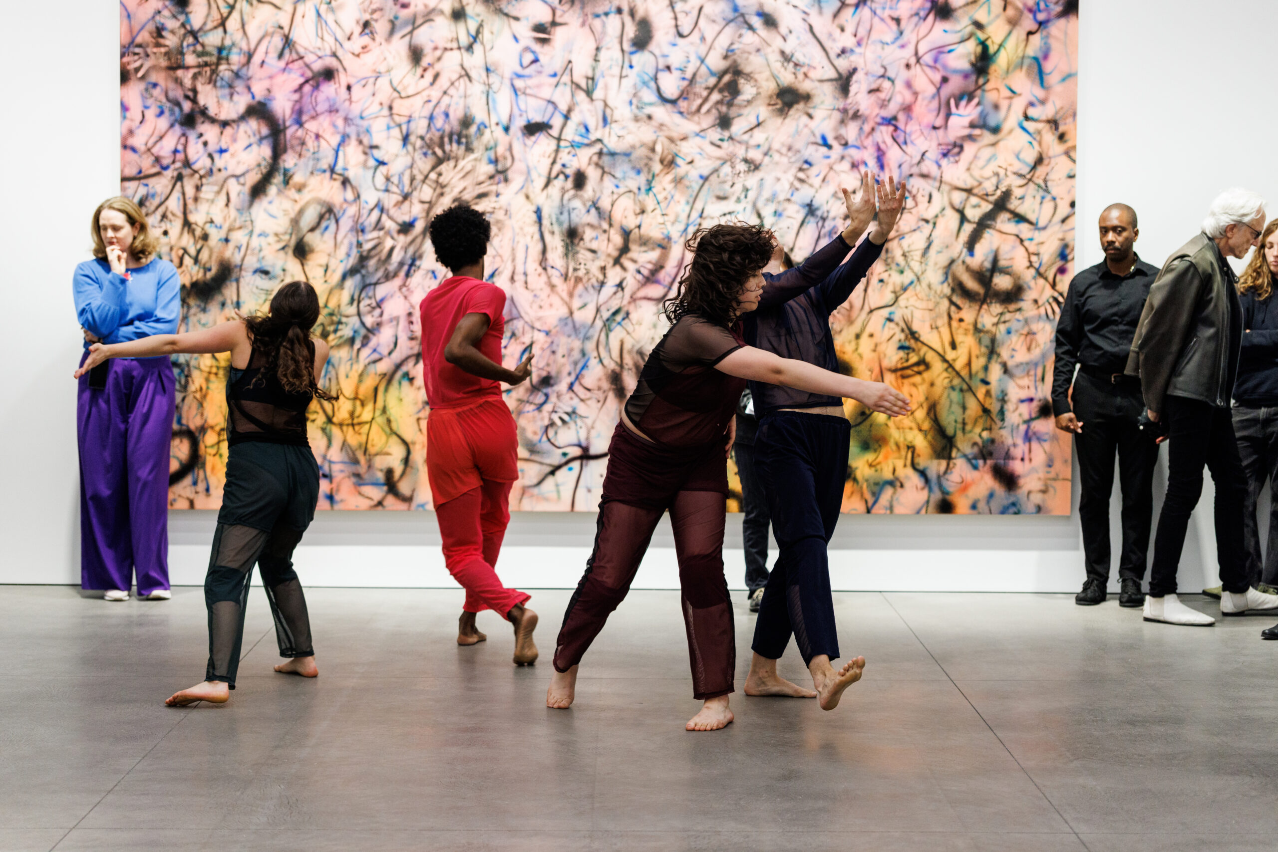 Four dancers move in front of a large abstract painting rich with textured color, their bodies reaching, extending, and twisting in multiple directions. Their hands and feet reach upwaerds, downwards, and away from us, creating dynamism in the air. Along the edges of the painting, bystanders stand and look--some at the dancers, others at something unseen.