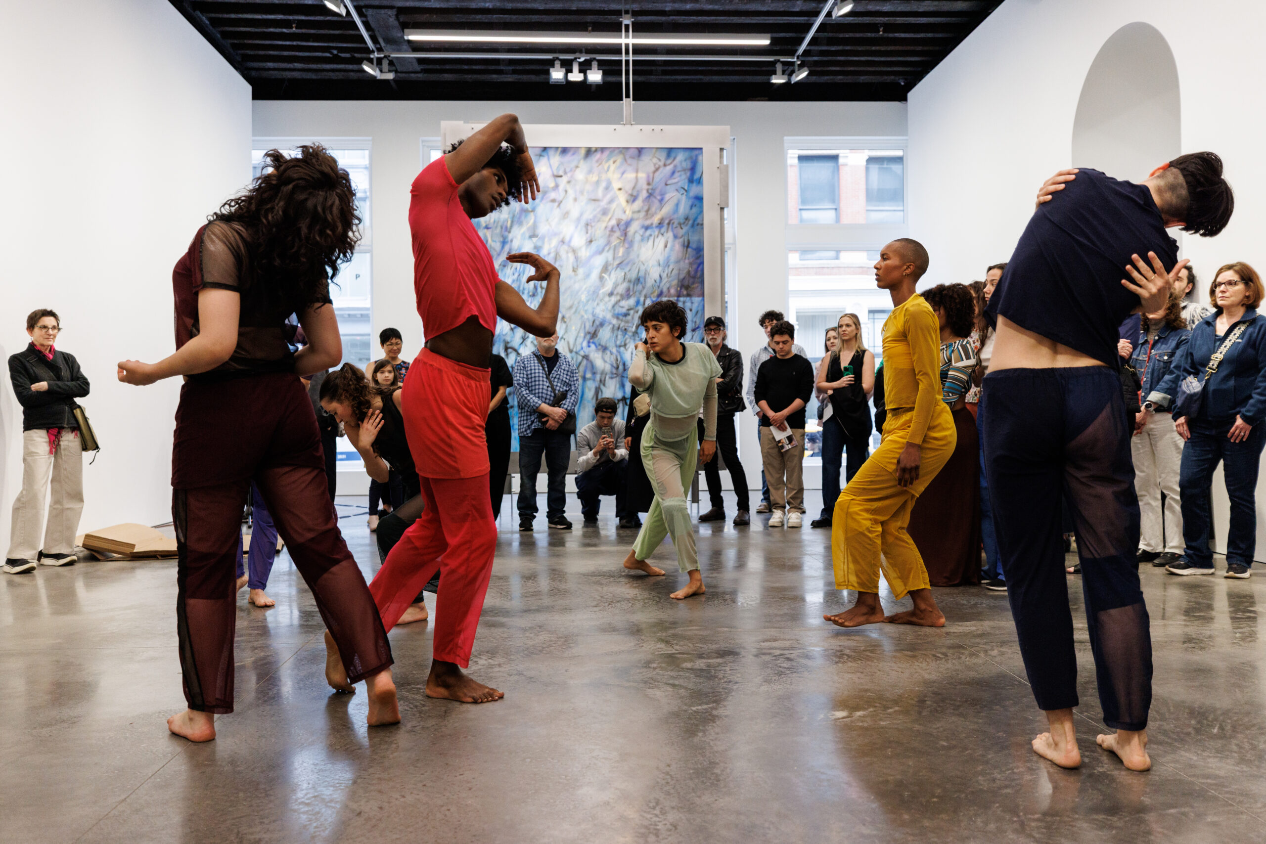 Six dancers are captured mid-motion in an open gallery space, a large blue abstract painting hanging behind them in the distance, and with about twenty people forming a half-circle around them. The dancers wear various costumes of a solid color--orange, black, red, gray--and all face different directions, moving in individual ways. One hugs themself, one twists their upper body to the side; one arches their chest upwards, another stands in profile with bent knees. The audience of all ages stands around them attentitively, some holding programs, cameras, or phones.