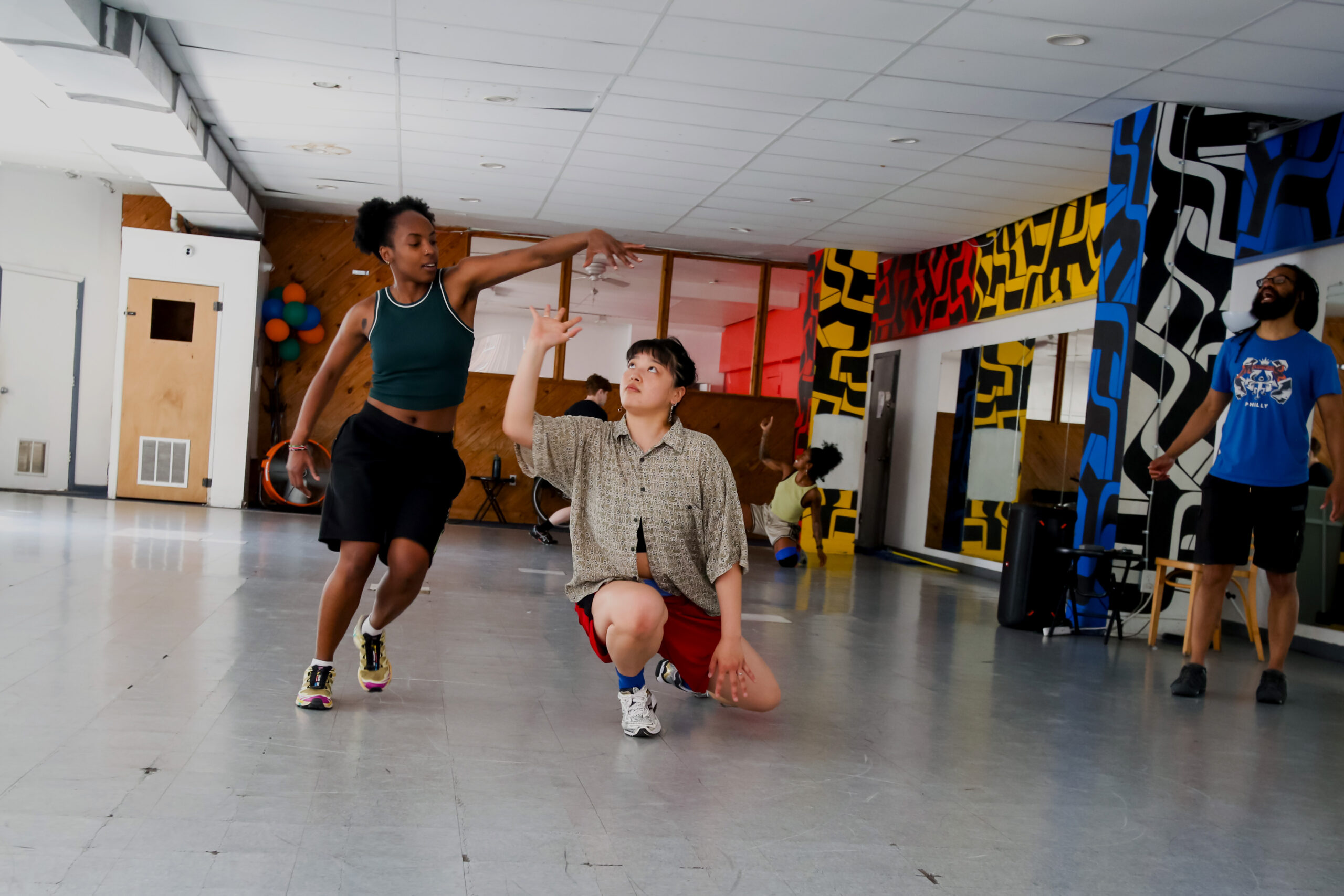 A spacious dance studio with a gray floor, mirrored walls, and colorful geometric murals is shown during a rehearsal for First Floor Spectrum. In the foreground, two people interact through expressive movement: one stands with an arm extended overhead while the other kneels and reaches upward toward the raised hand. Additional people are visible in the background practicing choreography, while another person stands near the right side of the room, directing. The studio contains chairs, exercise equipment, and a cluster of colorful balloons near the back wall. Natural light enters from windows along the left side of the space.