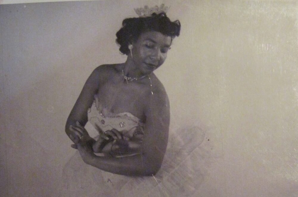 Black and white photo of Sydney King posing in a tutu and matching crown. Her arms are gently crossed in front of her torso and her head tilts back as if looking over her shoulder. King wears point shoes and stands in tendu derriere, one leg extended behind her.