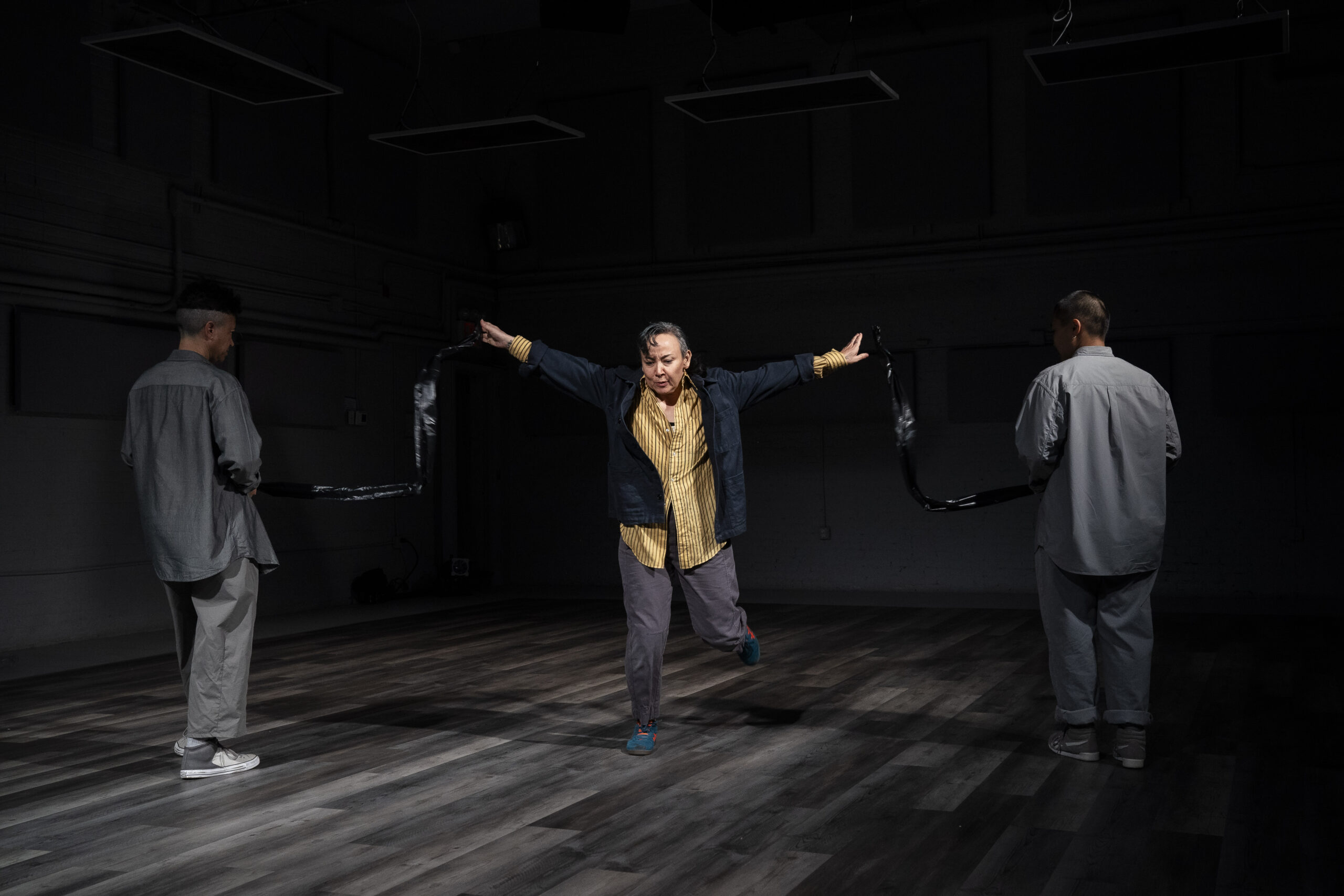 maura nguyễn donohue lunges forward onto one foot with her arms slicing outwards from her back. She wears a mustard yellow button-down shirt, navy blue coat, grey pants, and vibrant blue sneakers. She is framed by the grey-shirted backs of Shannon Yu and Rami Margron, and the darkness behind her.