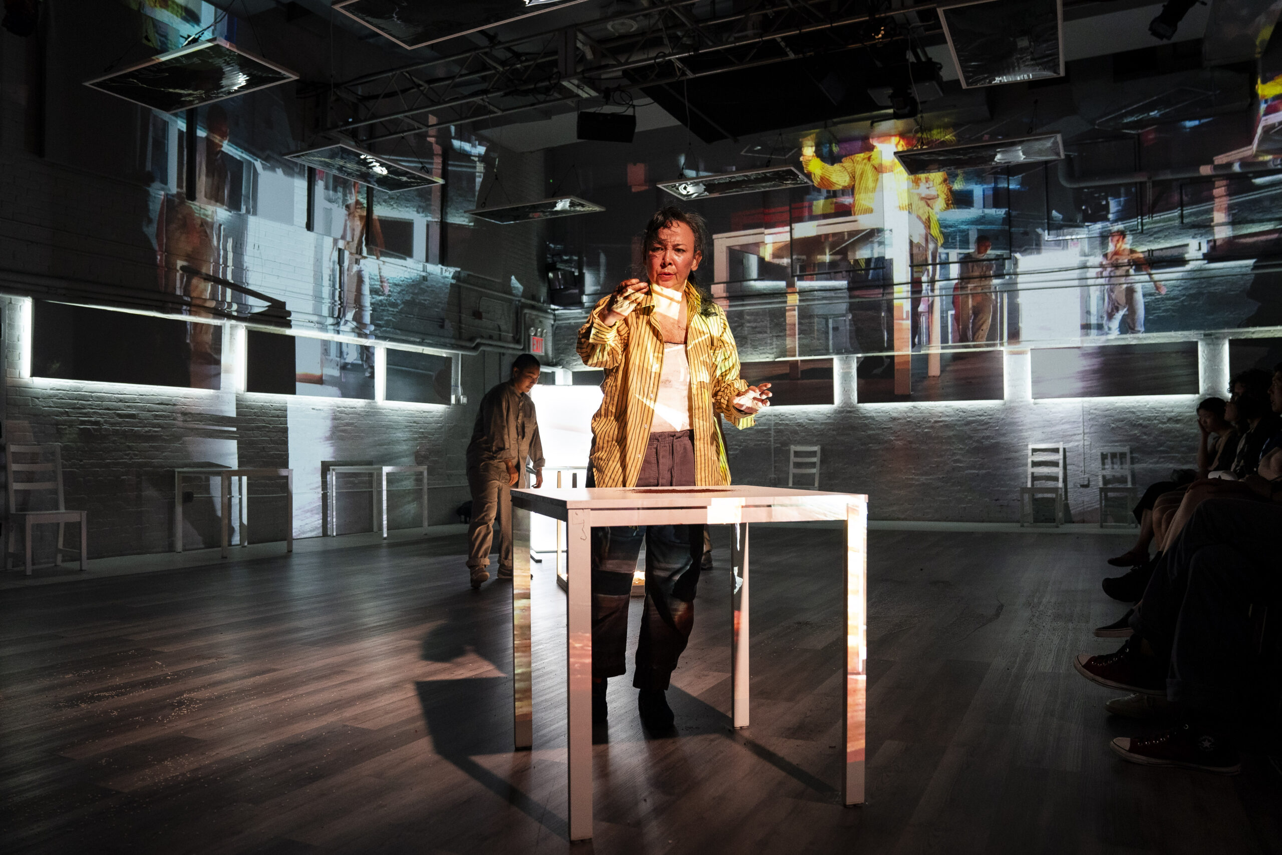 maura nguyễn donohue stands behind a white table, with overlapping projections of herself and the room around her covering her face and body. She wears a mustard yellow button-down shirt and holds a small object out in front of her, looking at it with curiosity. Shannon Yu is standing, slightly slumped, behind nguyễn donohue. Yu wears a monochrome grey outfit, and gently shrugs her head and shoulders forward, curving her upper spine.