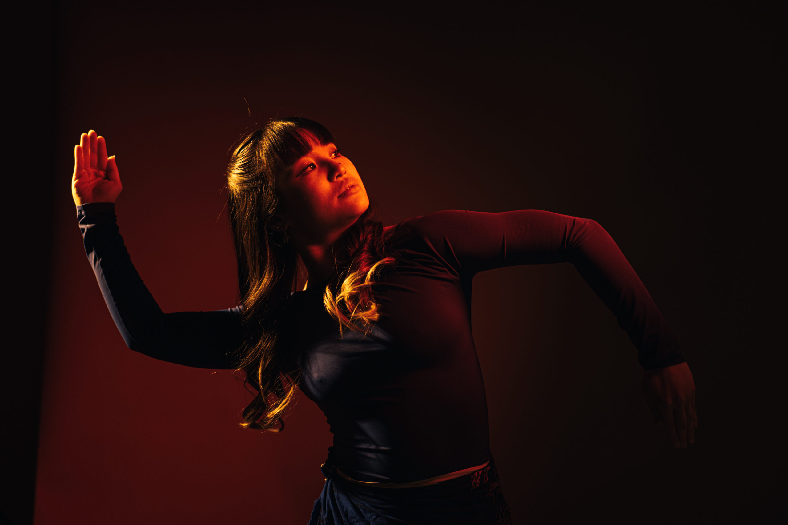 Jazlyn Tan, clad in a long-sleeve black top, leans her weight to one side. Her hands are blades; one arm in at a ninety-degree angle up, the other an inversion. Her gaze is focused and serious.