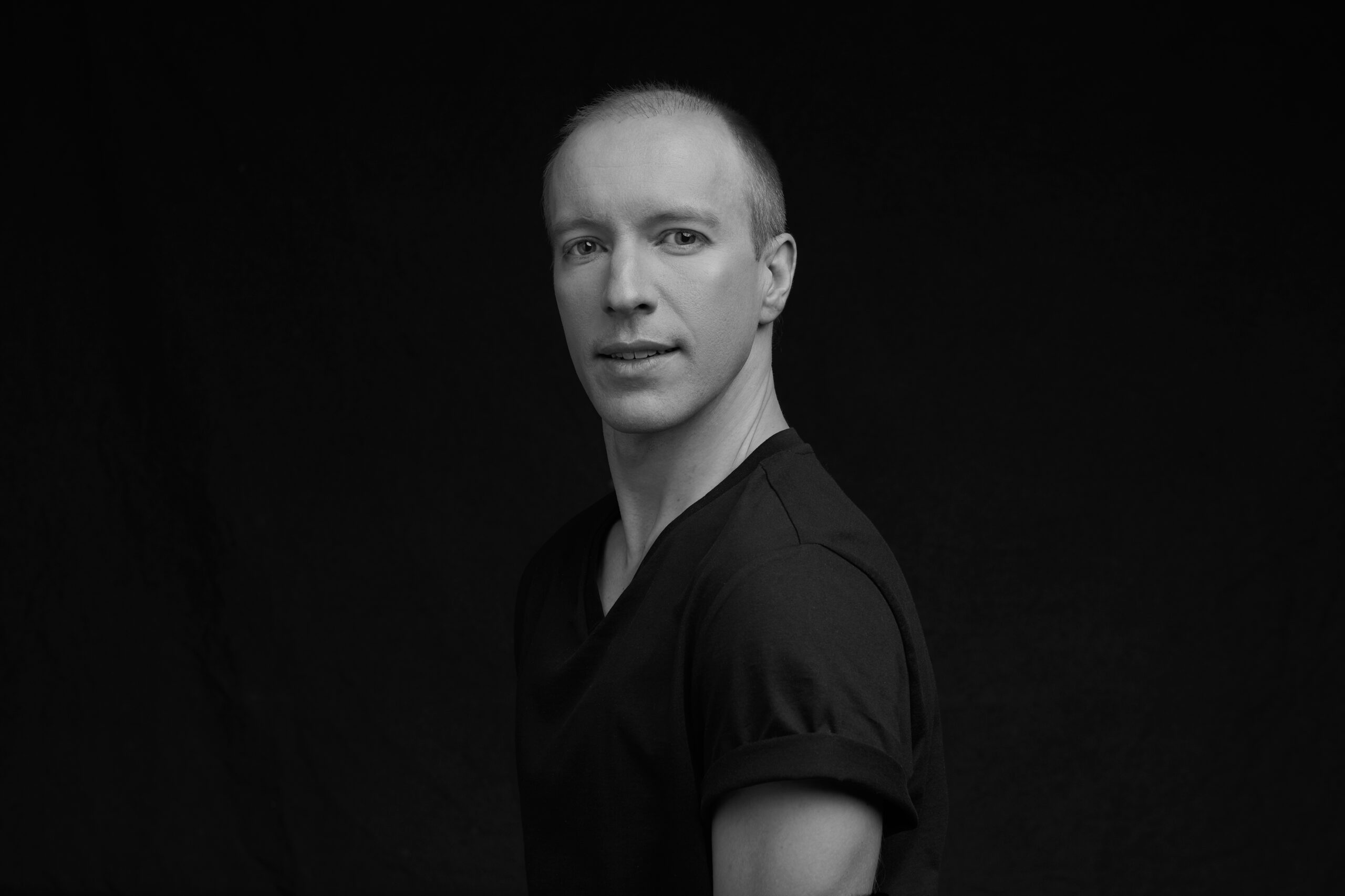 Andrew Skeels, a White man with buzzed hair, wearing a black T-shirt, stands against a black background. He is turned to the side but looks directly out with a soft smile.