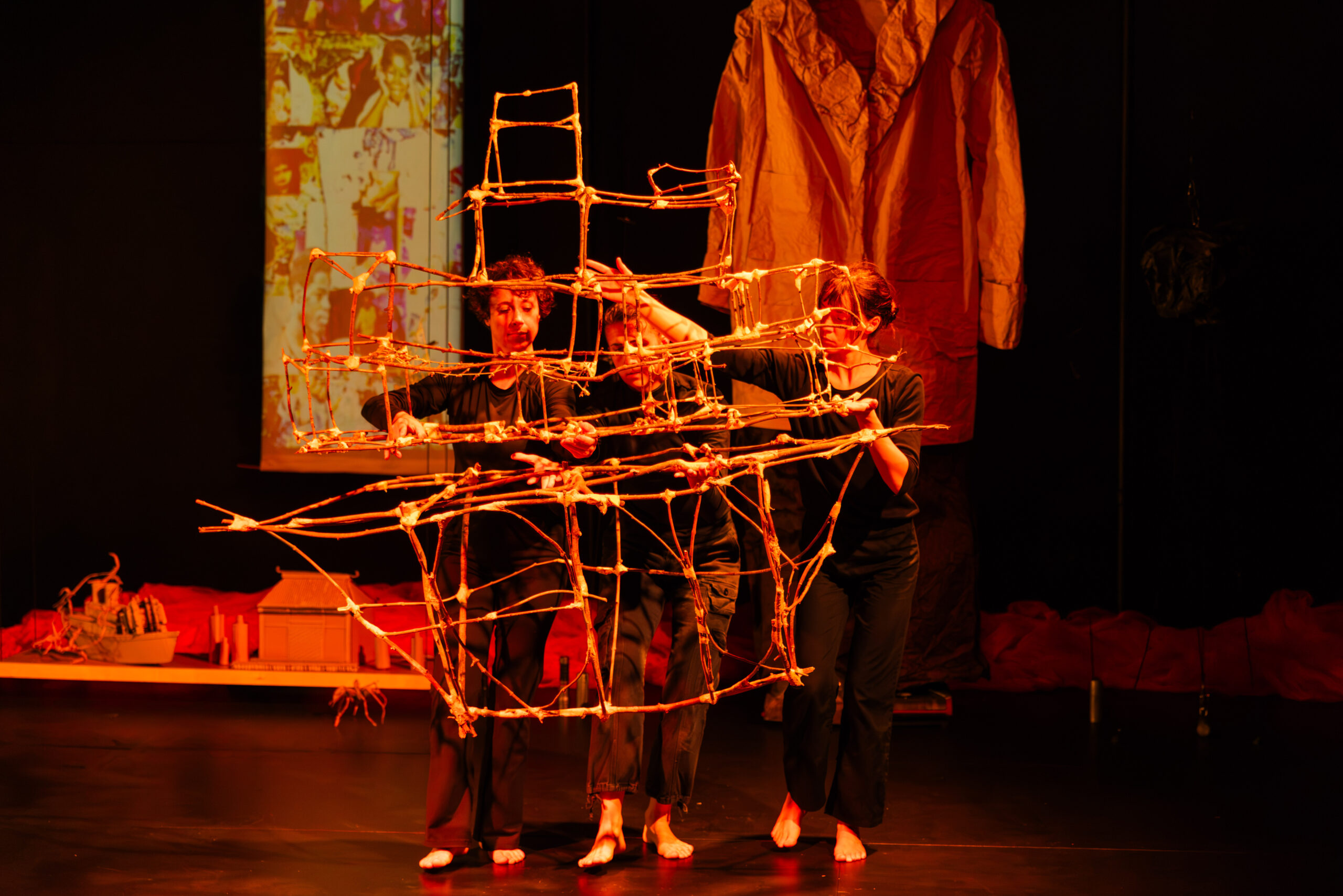 Awash in a red light, three femme presenting performers dressed in black hold a boat constructed out of tape and twigs. They are putting two halves of it together: one looks like several apartment buildings all together and the bottom is a clearer boat shape. An oversized coat hangs in the background, along with many different puppets from previous moments scattered along the back of the stage.