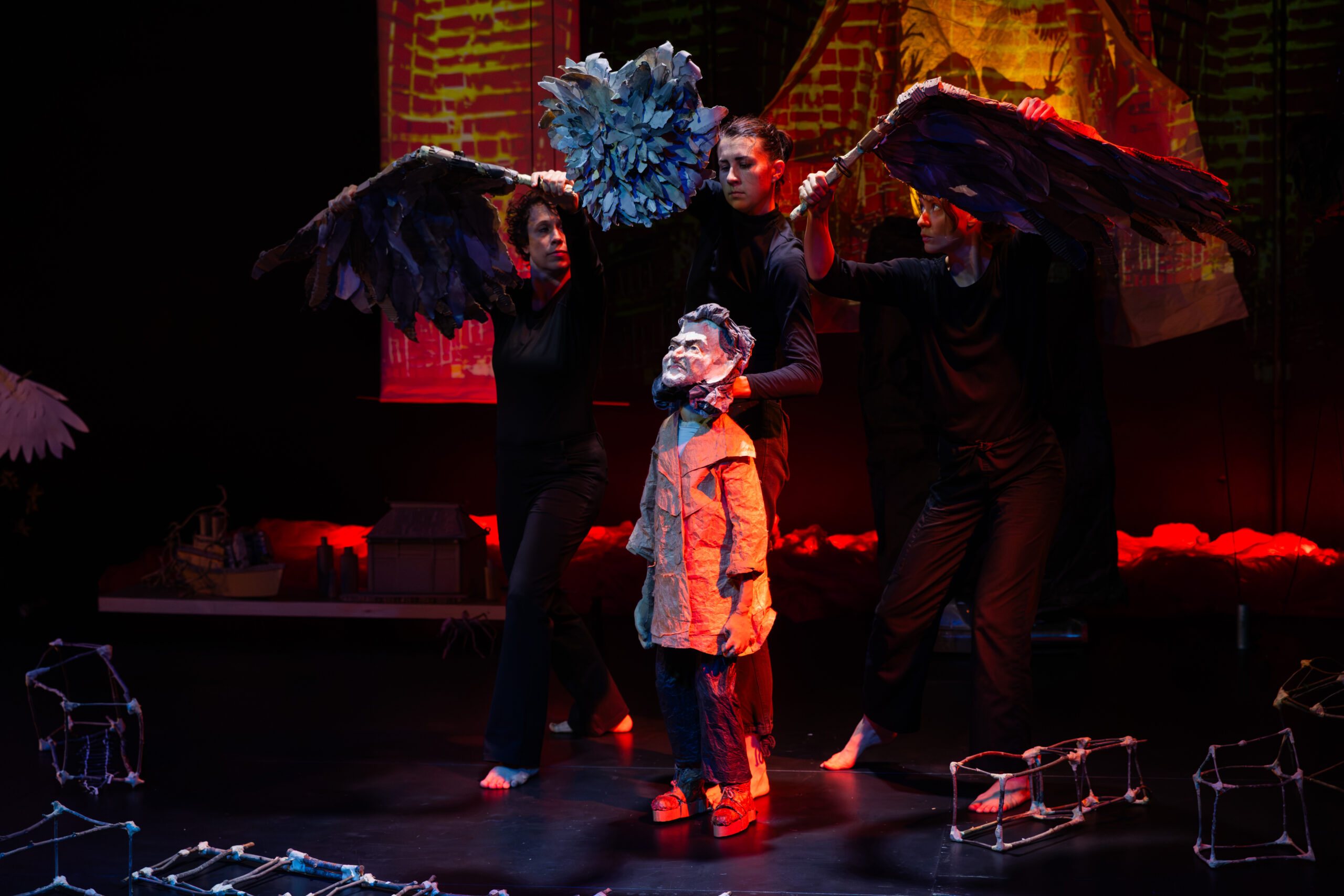 A papier-mache puppet of Kea Tawana is overshadowed by two massive disconnected owl wings and an eyeless owl head soaring above her, held by the puppetering cast dressed in all black. The Kea puppet is wearing an oversized trench coat, trousers and sandals. Parts of her ark lie scattered around her, made of twigs and tape. There is a spotlight over her, and the rest of the stage is darkened.