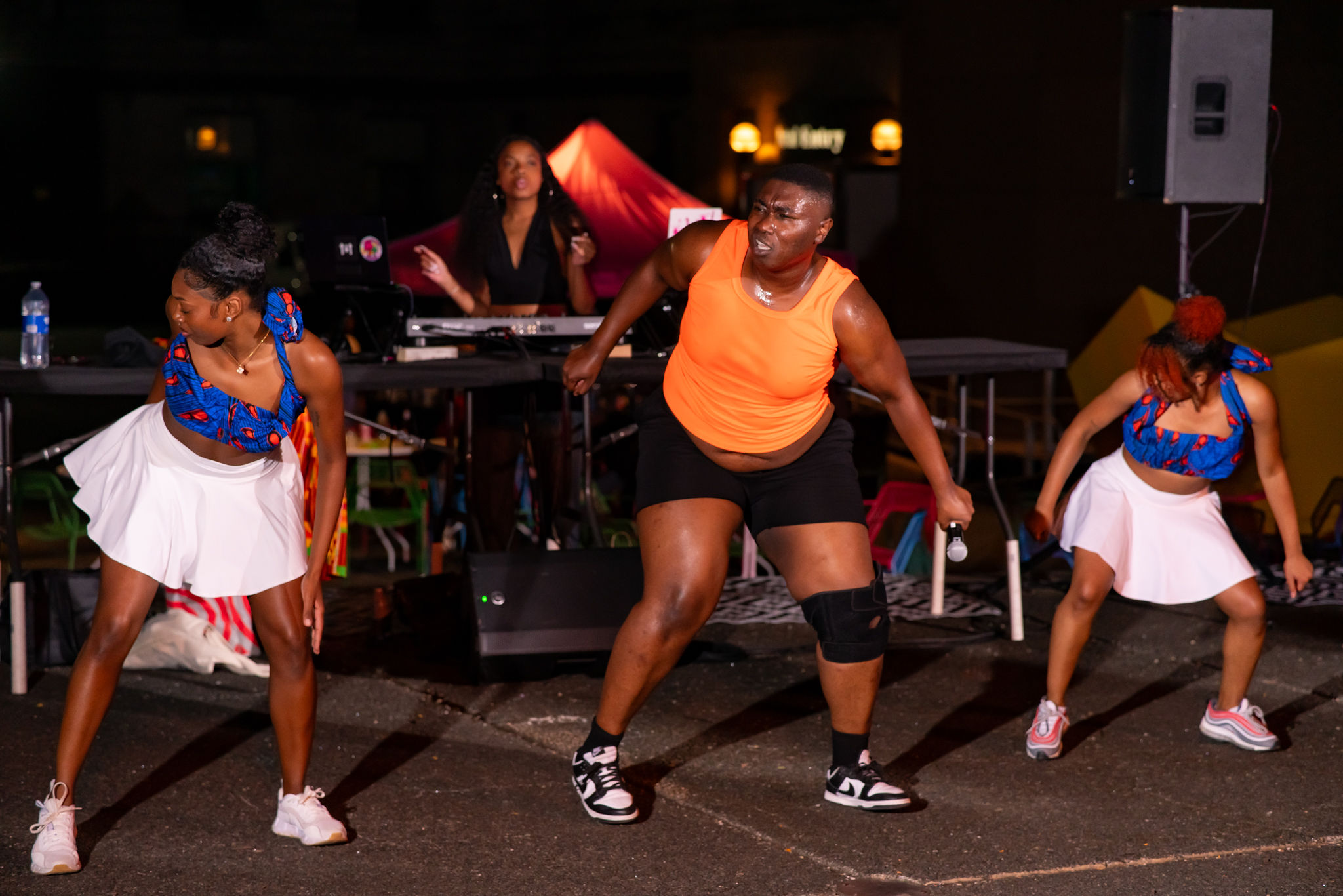 Bempa, wearing a bright orange shirt, black exercise shorts and sneakers, is framed by two black and brown femme dancers wearing matching blue and white outfits. They are dancing outside at night, lit by stage lights on a sidewalk with a femme appearing DJ playing music in behind them. The dancers have bent knees fronts facing the audience mid twist, looking over their shoulders with concentrated expressions.