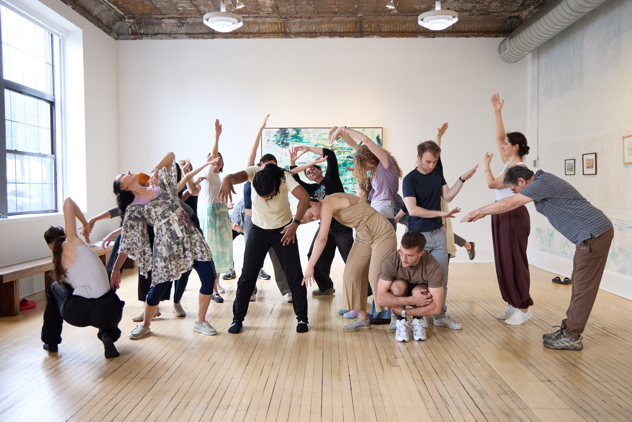 Around 20 workshop participants pose in front of an abstract oil painting in a bright gallery. They are all in different positions inspired by the artwork. Many stretch their arms out and up as if reaching. Some arch their backs or bend forward.