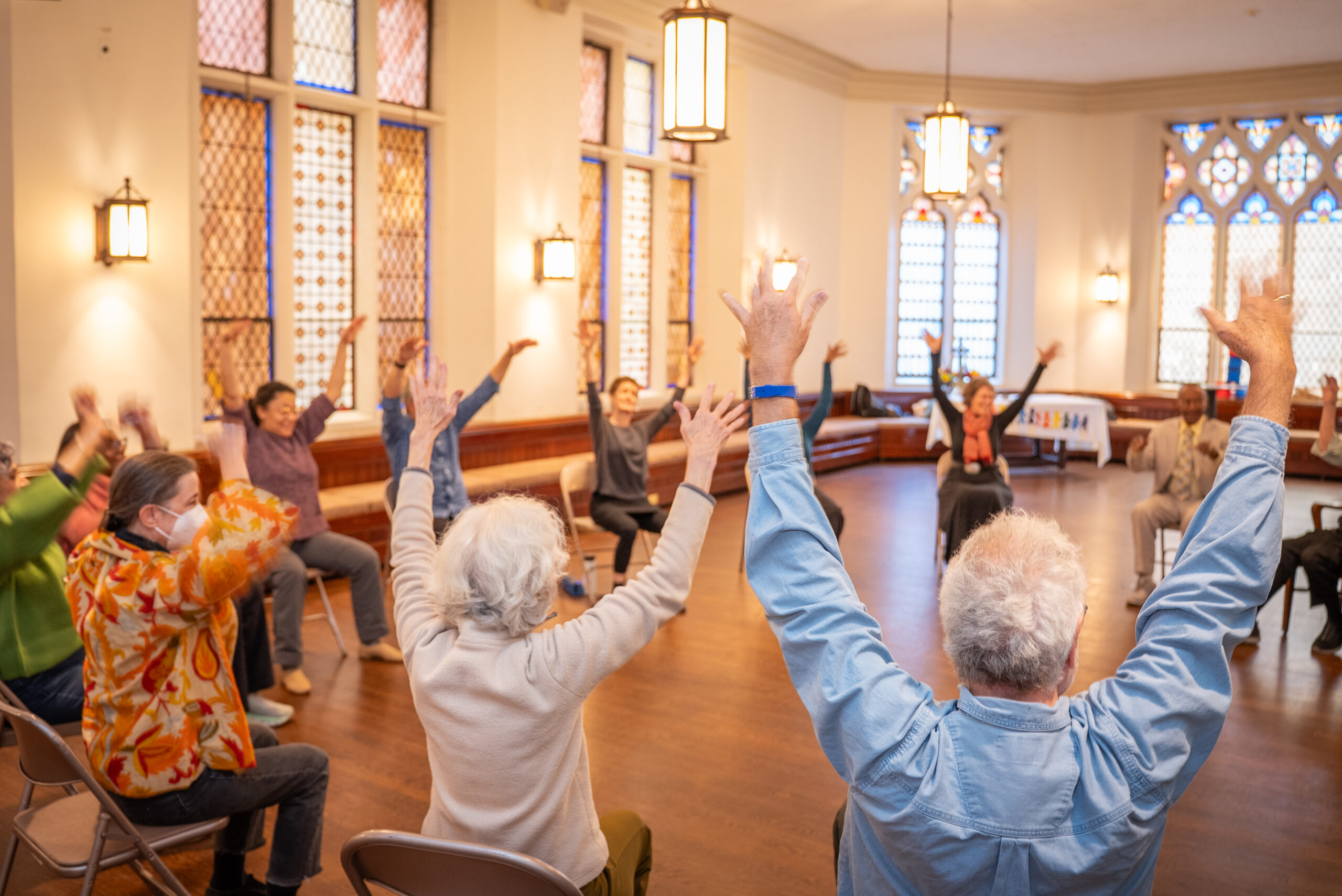 A group of dancers wearing various multi-colored shirts sit in chairs arranged in a circle in the center of a church meeting room. They are surrounded by intricate stained glass windows, warm wooden flooring, and off-white painted walls. As they sit, they raise their arms above their heads and smile at one another.