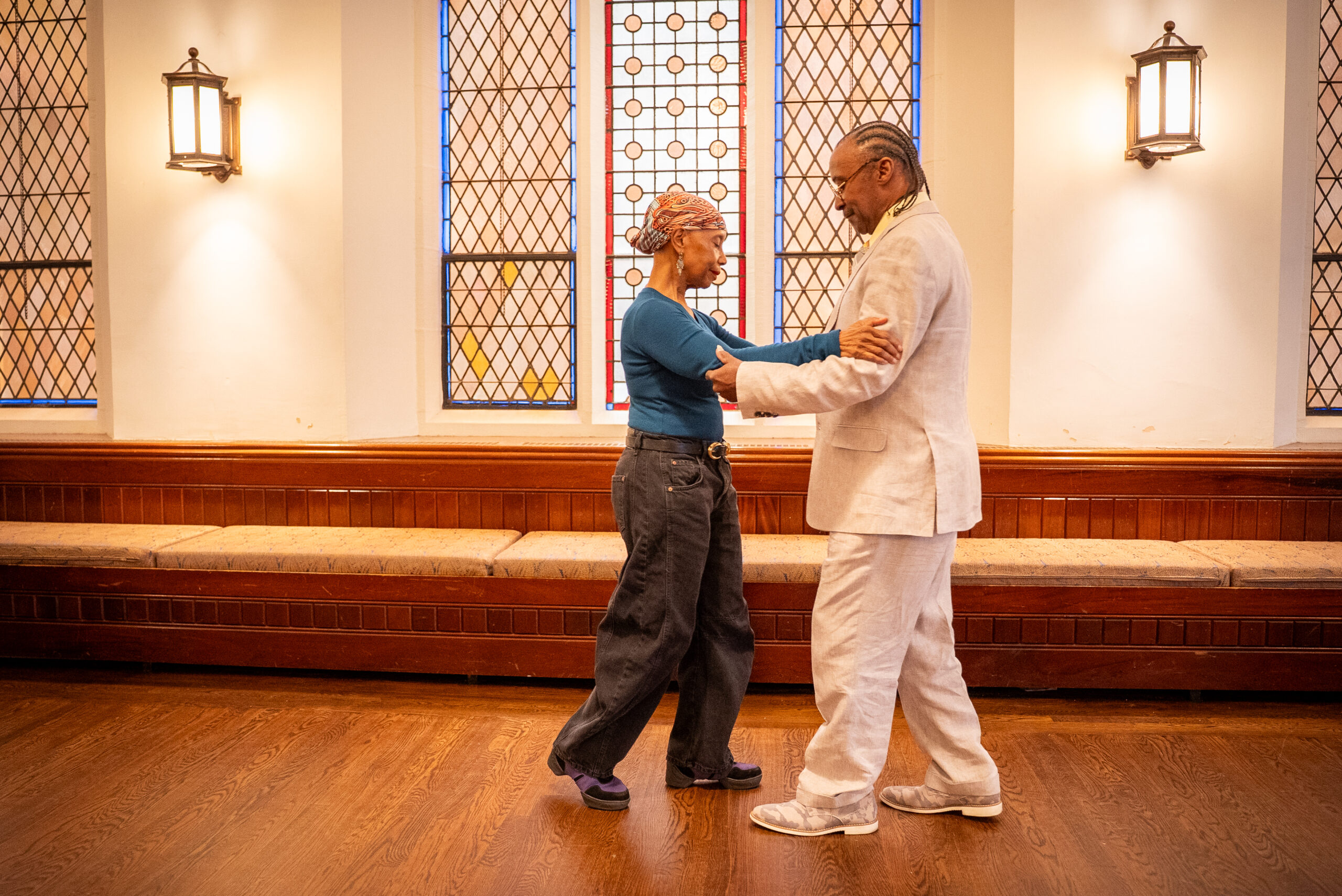 Two dancers stand facing one another, gently resting their hands on the other’s forearms. As one dancer steps forward, the other steps backward. The leading dancer is a woman, who wears a brightly colored headscarf, dangling earrings, a vibrant blue top, and stylishly baggy black jeans. The second dancer is a man, wearing an off-white linen suit, with his hair braided into neck-length cornrows. Both dancers are glancing towards the other’s feet as they move in sync through a partnered Tango sequence.