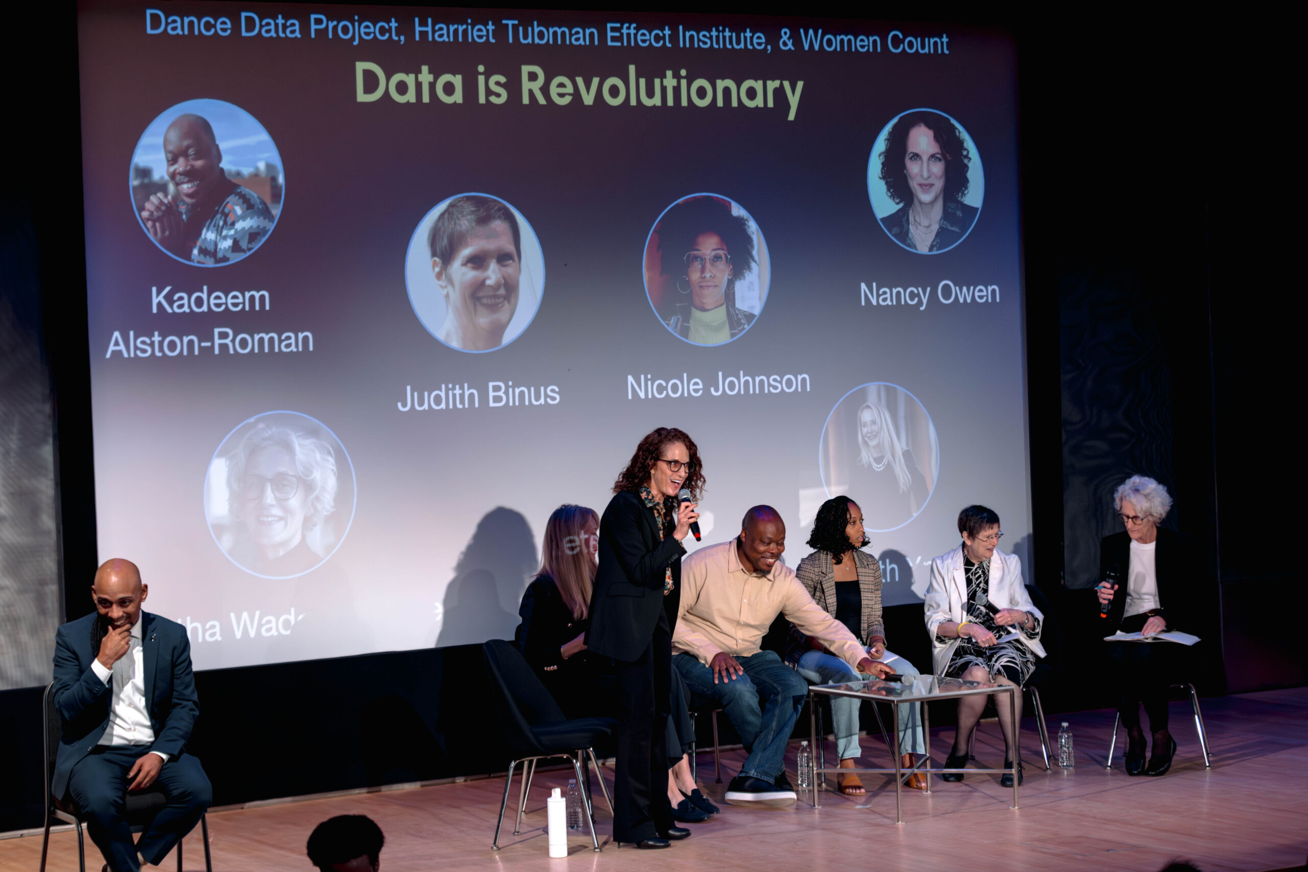 At the RISE 2025 Summit held at the New York Library for the Performing Arts, panelists for a discussion entitled “Data is Revolutionary” gather in black office chairs with silver legs in front of a large projector screen that reads “Dance Data Project, Harriet Tubman Effect Institute, & Women Count” in a pastel blue above the title “Data is Revolutionary” in a light yellow. Six circles containing the headshots of the panelists dot the rest of the projection. Amidst the other seated speakers, Nancy Owen stands holding a microphone in her right hand. She leans forward toward the audience, eyes alight, mouth open in speech that slips through an excited smile. In the lower left corner of the frame, a bald man in a crisp blue suit brings the crook of his thumb and second finger to his chin at stage right. He looks out toward the audience with his chair angled away from the six panelists.