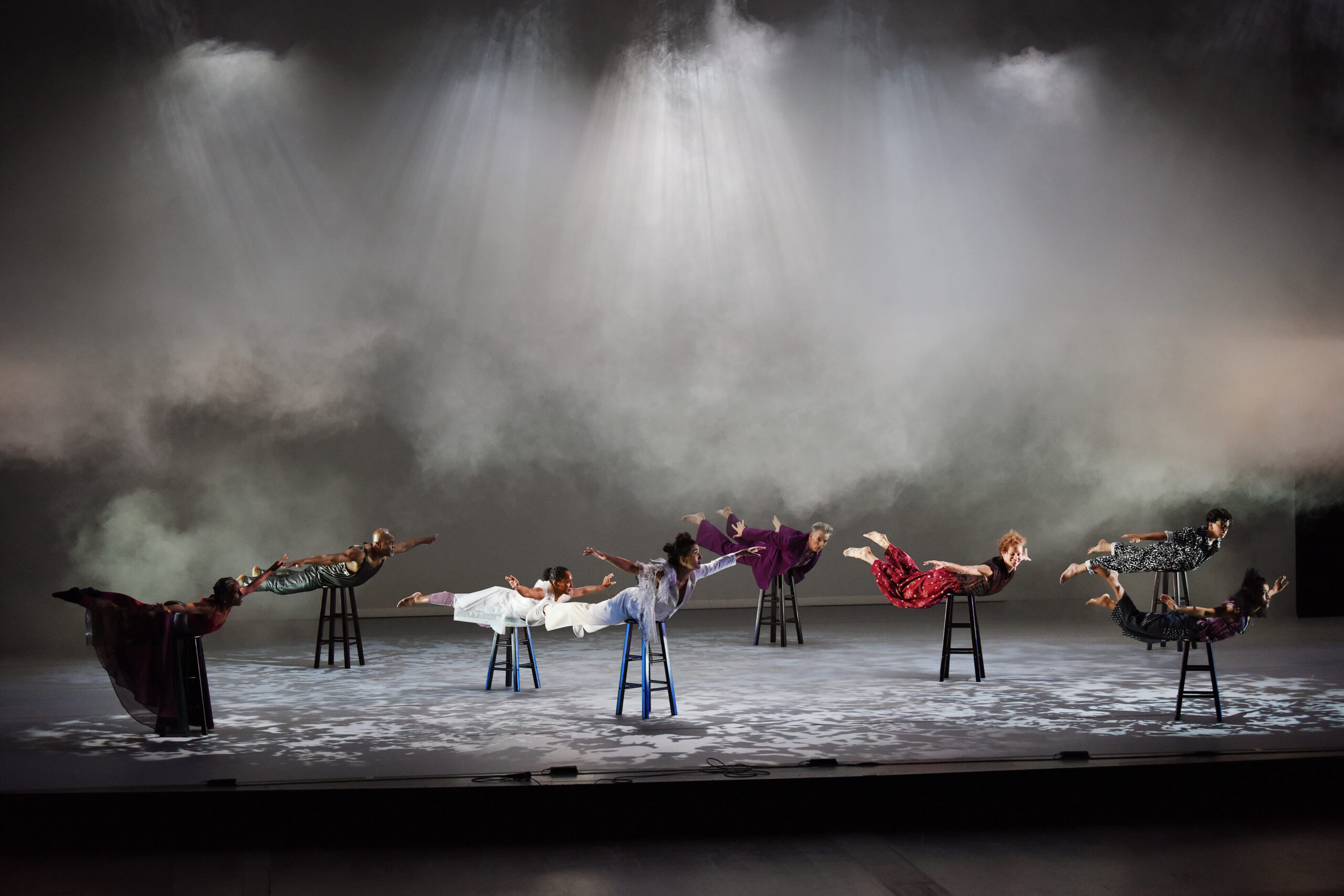 Nia Love, Vincent Thomas, Miko Doi-Smith, Paloma McGregor, Will Bond, Leah Cox, Elisa García Radcliffe, and Ruby Morales in Wicked Bodies at Arizona State University, Gammage (2022). Organized in three staggered lines atop individual black stools, (three dancers, two dancers, three dancers), the performers each assume a “superman” position on their stomachs, back muscles taught as arms and legs lift off. The dancers wear an array of costumes (billowing pants, structured blazer, expansive gauzy skirt, a polka dot jumper) in shades of black, white, fuchsia, and red. Collectively, they cast their gaze stage left, the direction of their “flight” as haze descends over the top ⅔ of the stage height.