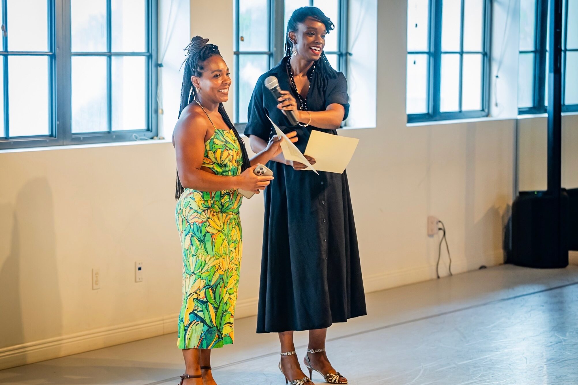 The co-founders of Banyan Dance Festival are two black women, Branndi Lewis and Ericka Squire. They stand in a dance studio, wearing cocktail attire, a green fitted dress for Lewis and a black button-down collared dress for Squire. With glittering smiles, they share a microphone and hold paper notes for their address to the intimate audience gathered at Ballet Florida for the Banyan Dance Festival gala and preview evening taking place the night before the mainstage performance.