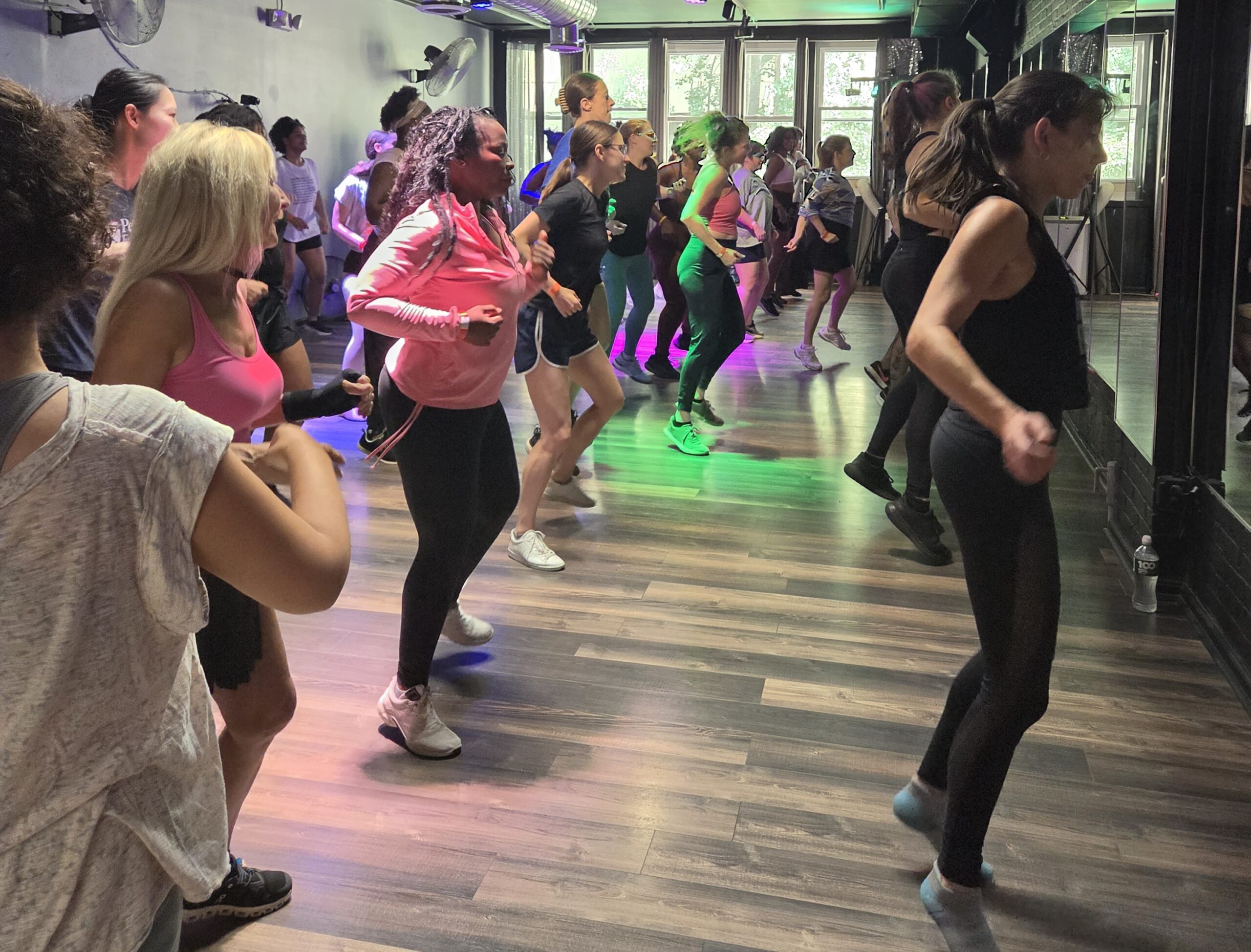 Several lines of students are mid bounce/step, focused on executing the choreography. All face front toward a wall lined with mirrors. The instructor stands in the middle of the front row of dancers. Participants wear a variety of athletic wear–workout sweats, tanks, shorts, long-sleeve zip ups–and most dance in sneakers.