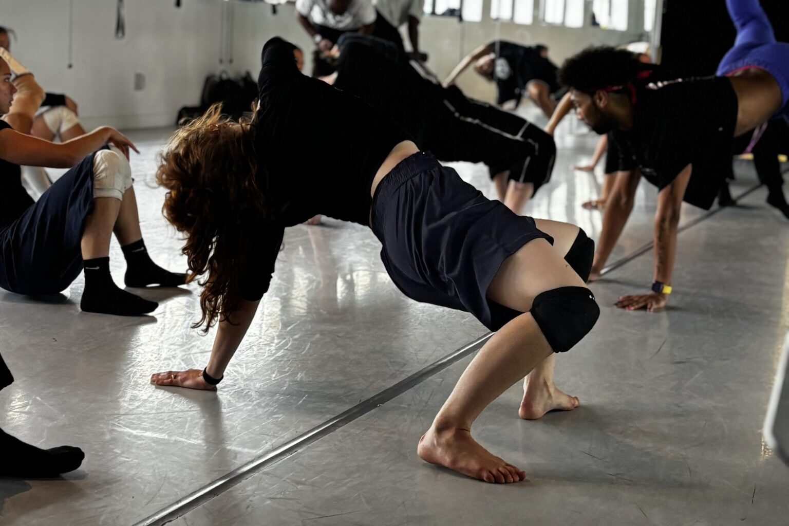 In rehearsal with Sean Aaron Carmon, the Resident Choreographer at Banyan Dance Festival’s Inaugural week, dancers practice phrasework close to a light grey marley floor. In the foreground, a young white woman with loose brown hair in navy basketball shorts, black kneepads and black socks hinges backward. Carmon, a young black man with curly hair and a red bandana demonstrates floorwork in the background of the photo. Other dancers are busy at work in the rest of the photo. Some are part way out of frame.