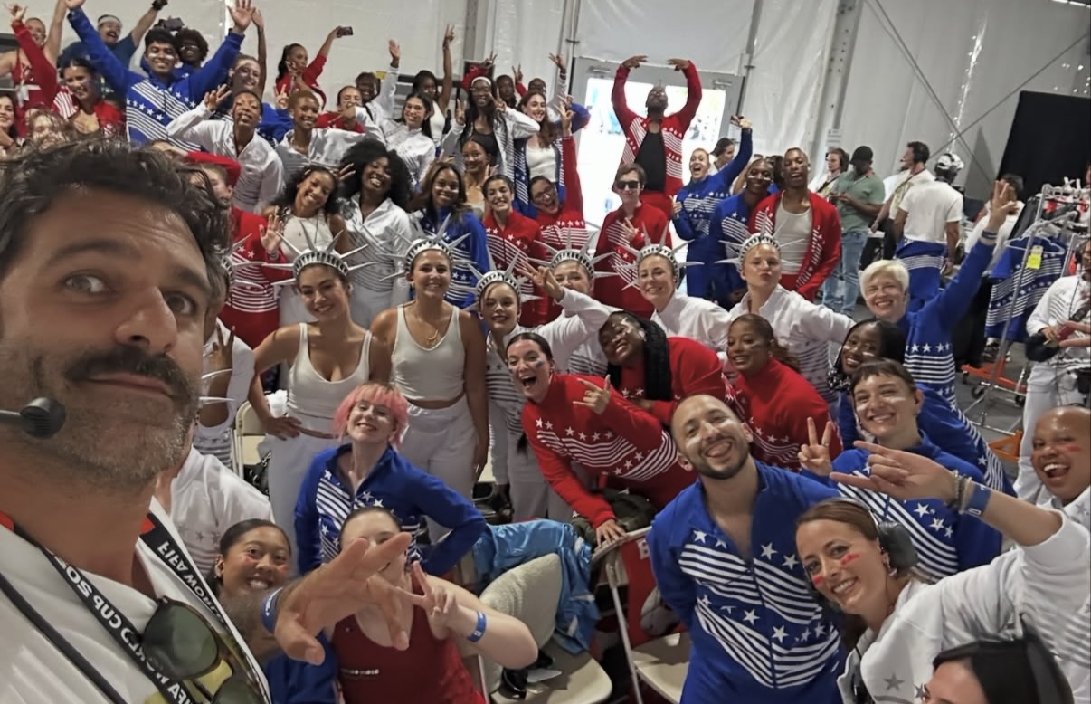 A wide-angle, high-energy backstage selfie featuring a large crowd of performers in a white tent. In the foreground, a man with a mustache and a headset takes the photo. Behind him, dozens of diverse performers smile, wave, and make hand signs. They are dressed in red, white, and blue tracksuits, with several wearing matching silver spiked Statue of Liberty crowns. Photo by Carlos Navarrete-Patiño, Production Designer. Production by Balich Wonder Studio.