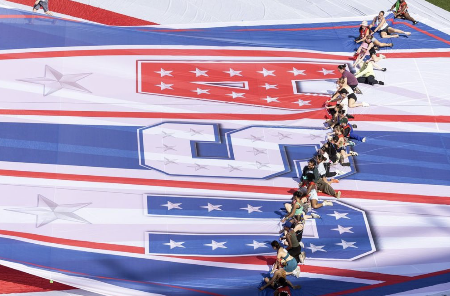 An overhead, high-angle view of a long line of people working together to unroll or pull a massive, vibrant patriotic tarp across a white field. The large-scale fabric features the letters "USA" written sideways in bold block lettering decorated with stars and stripes. The letter 'U' is blue, the 'S' is white, and the 'A' is red. Photo credit: Balich Wonder Studio.