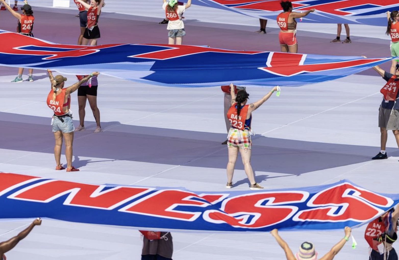 An overhead shot of performers on a massive white stage or field holding up giant, elongated banners. The banners feature large red text outlined in white against a blue background, spelling out excerpts like "HAPPINESS" and "LIFE, LIBERTY". The performers wear numbered red vests over casual clothes and hold their arms high to display the fabric. Photo credit: Balich Wonder Studio.