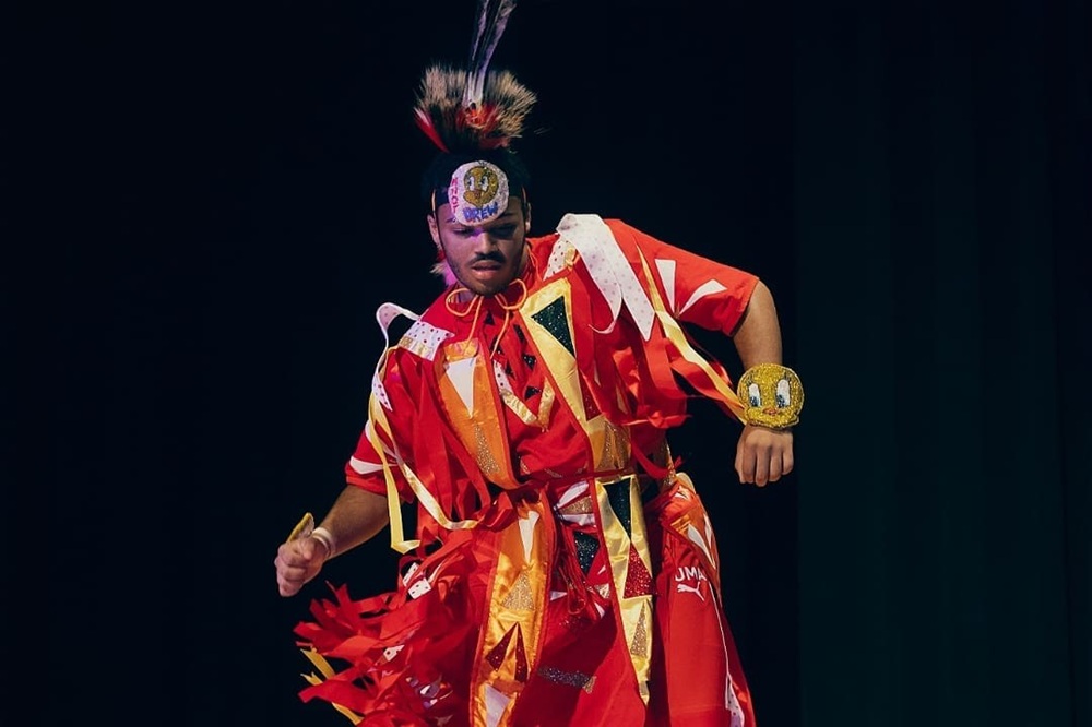 A dancer wears a feathered head piece and bright red garment with yellow, orange, black, and white strips of fabrics that dangle as they dance in front of a black backdrop.