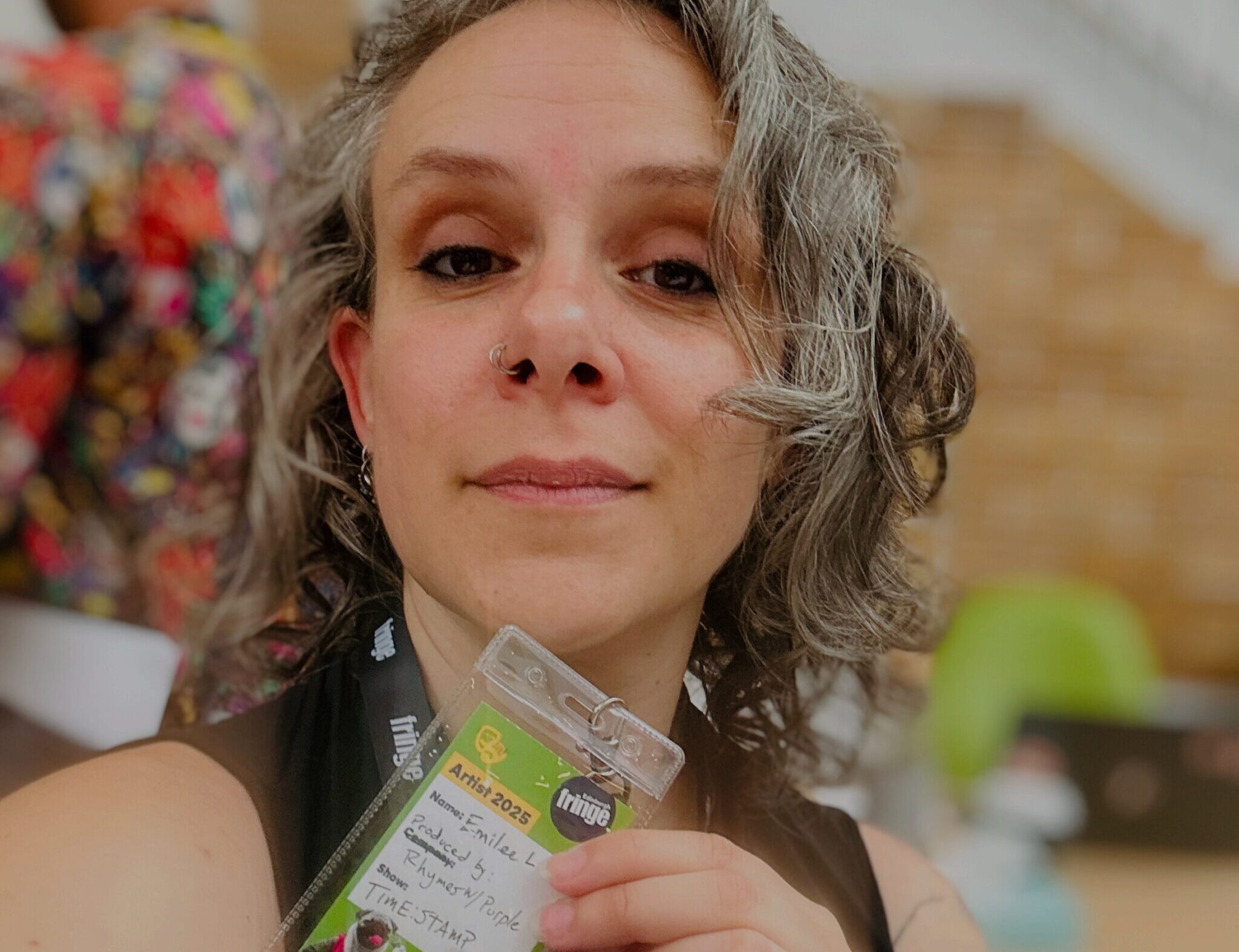 Emilee Lord, in a black tank top, smiles into the camera, her grey and brown curly hair framing her face. She holds the participant badge for Edinburgh Fringe in front of her chin.