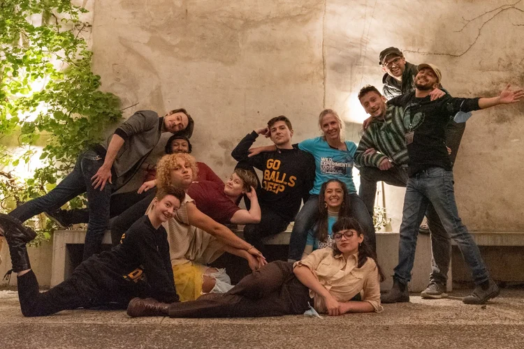 A group of twelve people pose in front of a white wall. on the floor level, four group members either sit, lie, kneel or lunge on the ground. standing members lean on one another on either side of the crew, and people in the center sit or lay on a bench together.