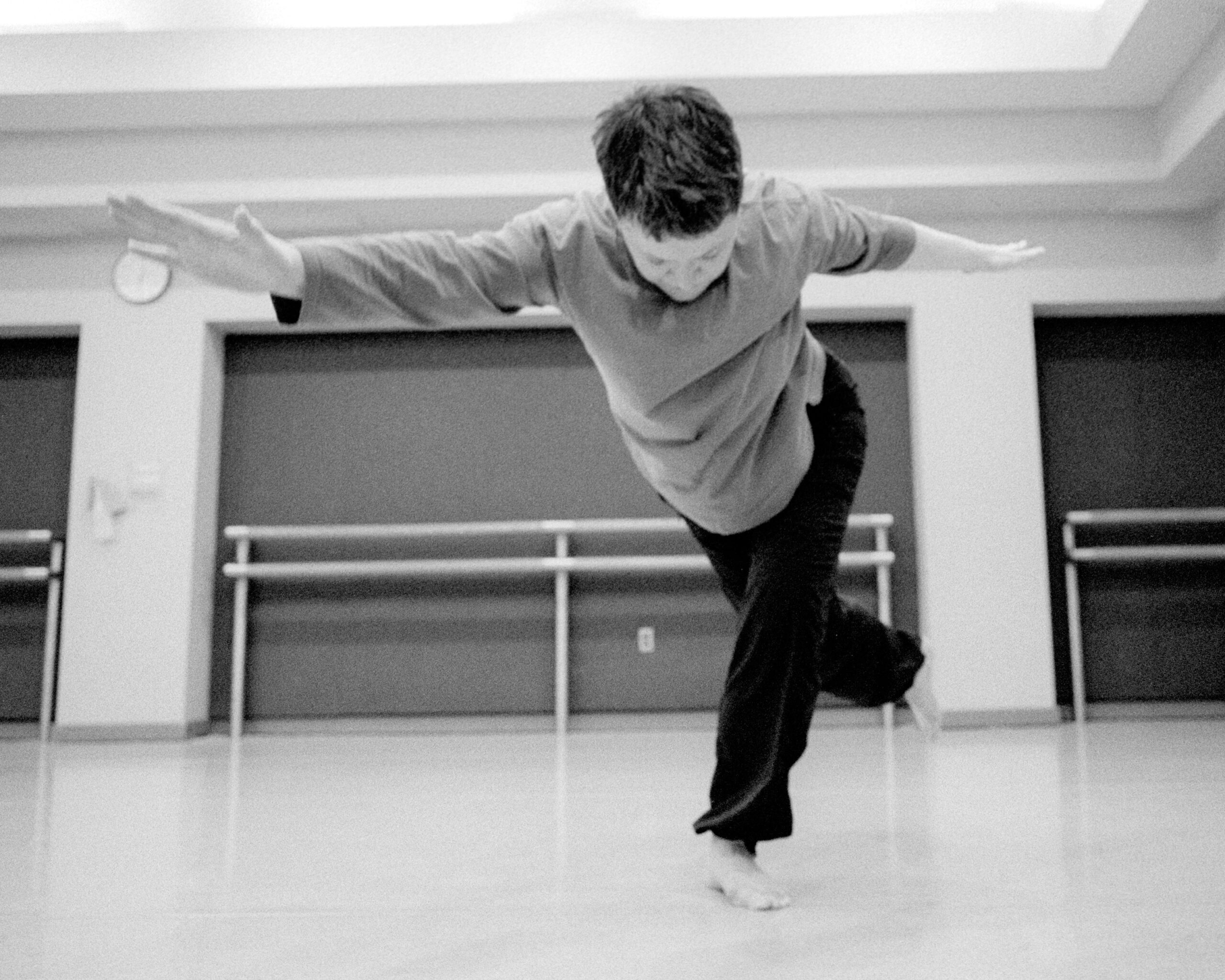 A dancer with short hair reaches their right arm forward, left arm back with their head bowed down. They stand with their left leg slightly turned out and bent, their right leg extended behind them.