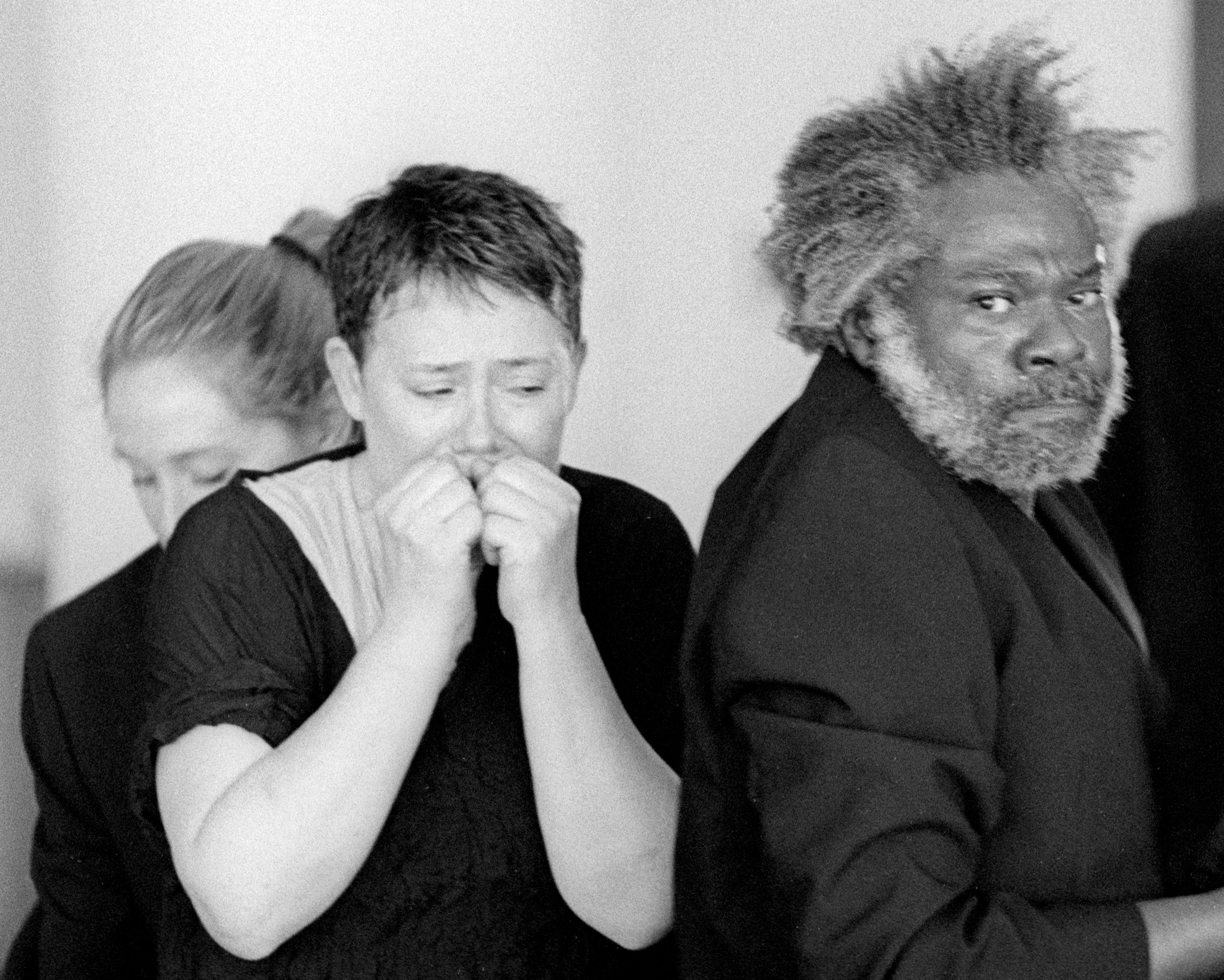 Three people are in this black and white image. The center person looks down, their hands in fists, scrunched up to their lips. Another man with a beard, wearing a suit jackets, stares directly into the camera. The last person is partially obscured, looking down.