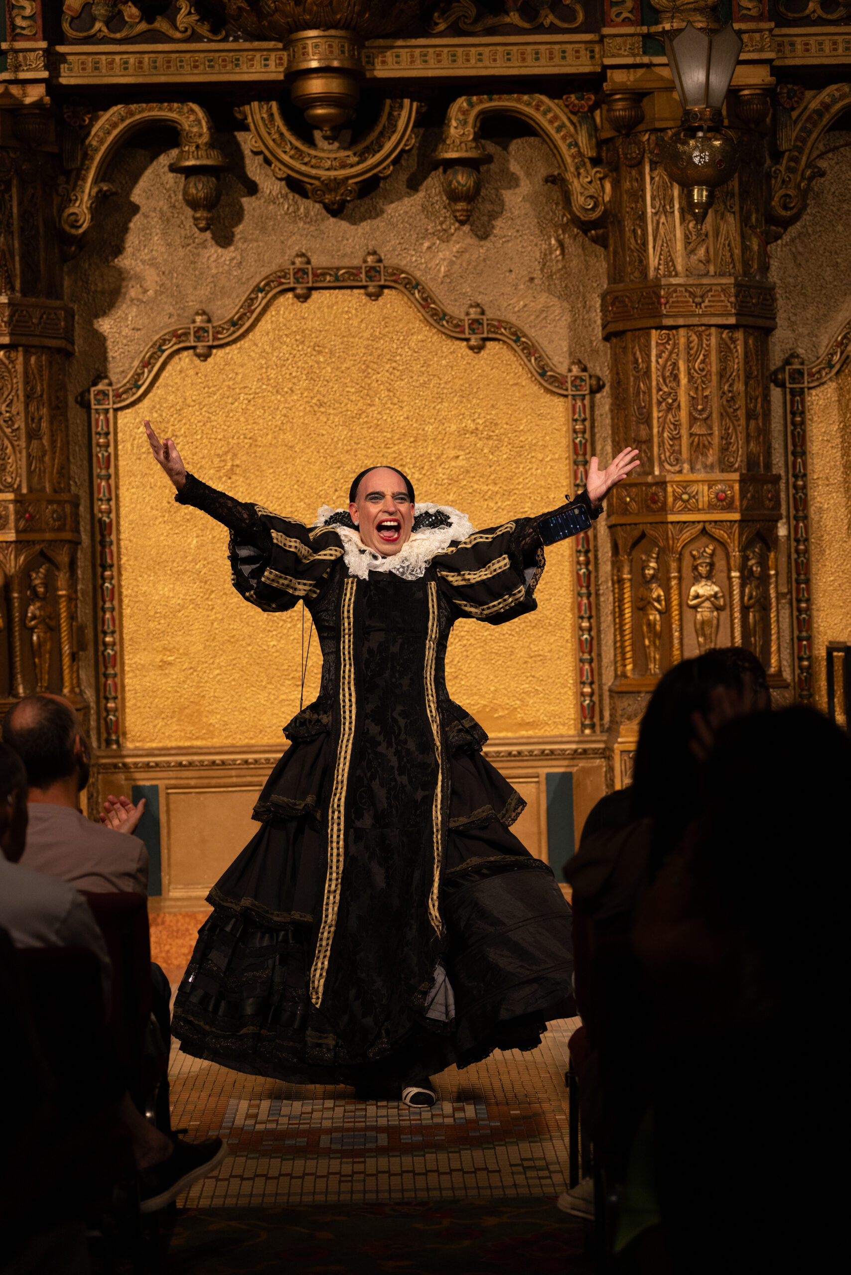 Edu Dìaz is dressed in a black gown with a large white lacy collar center on a golden gilded stage. Their arms are up exubrantly akimbo.
