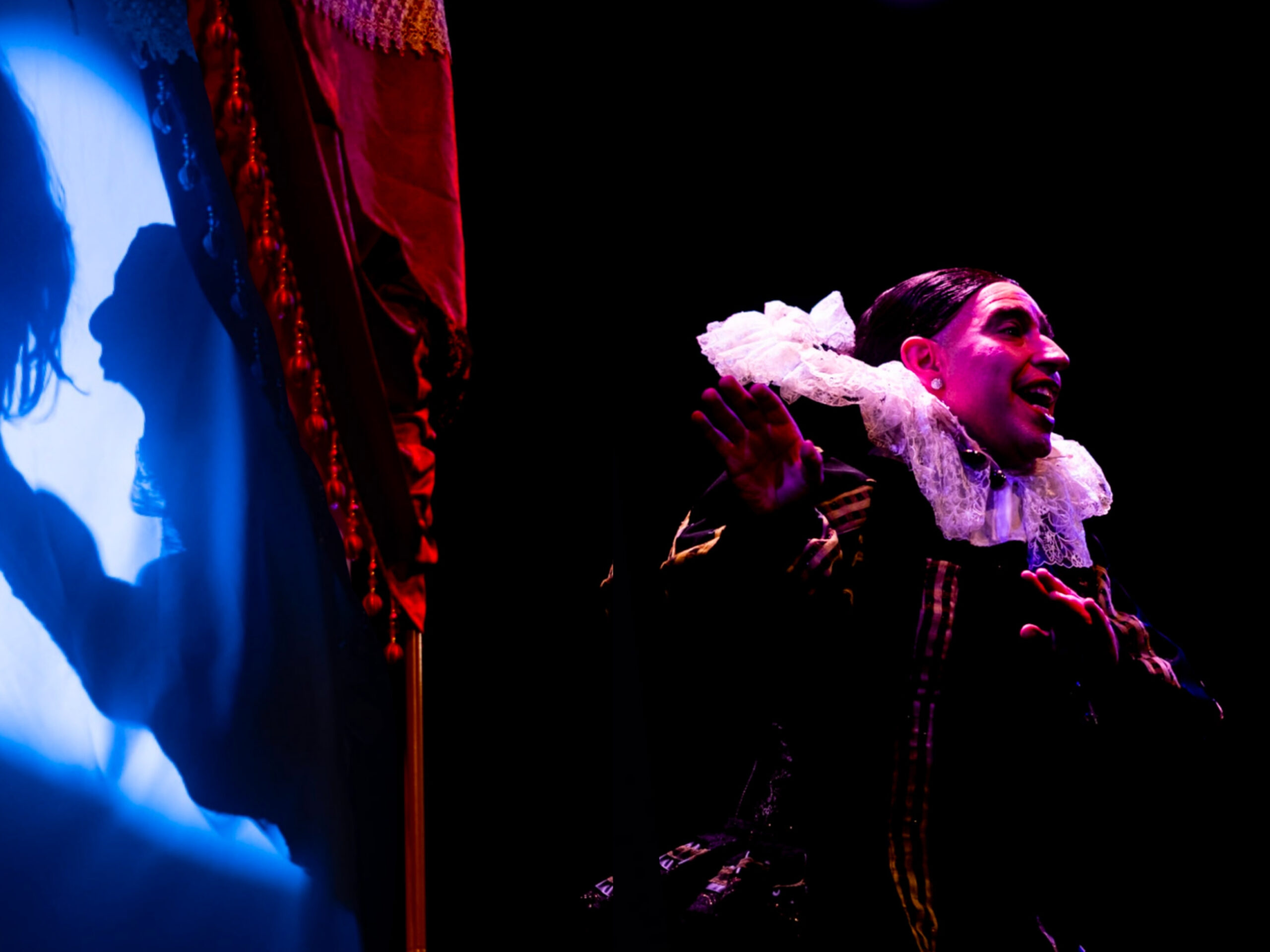 Edu Dìaz dressed in an ornate gown with a big white lacy collar stands speaking to the right. Behind them is a bluye projection of what appears to be someone screaming. The projection is framed by a red velvet curtain.