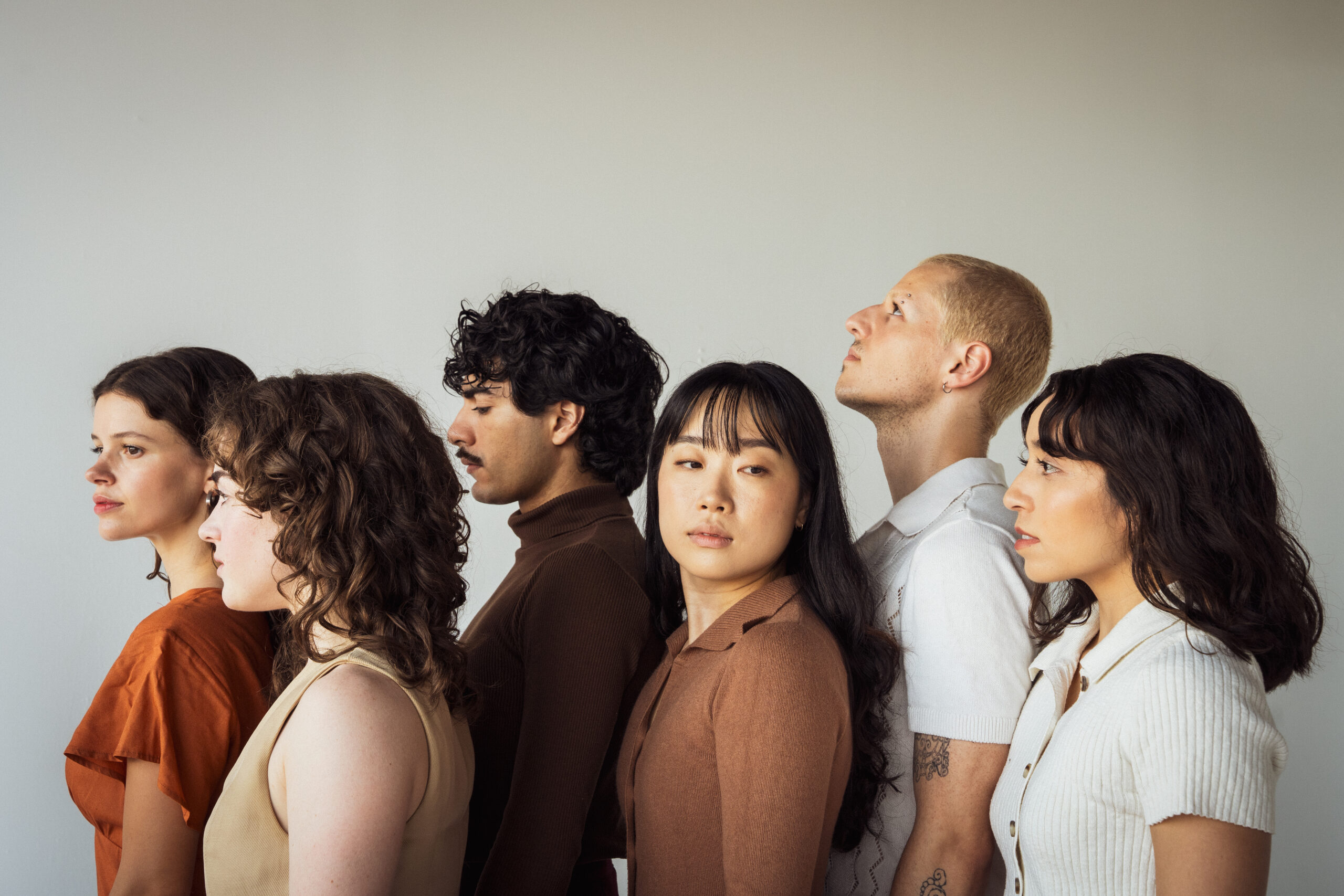 Five dancers stand in side profile, all looking in different directions and wearing earthy-colored cotton tops. Their faces are impassive and neutral.