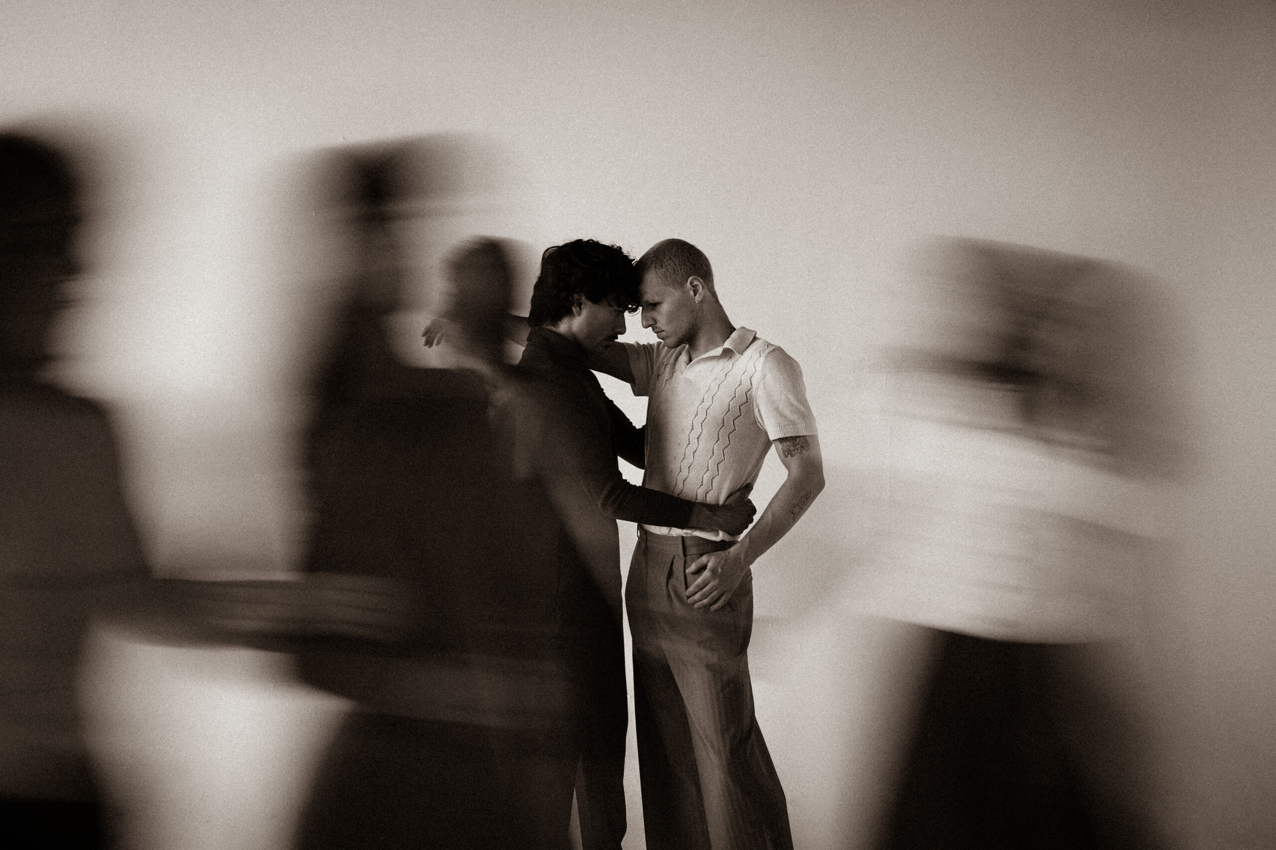 In a black and white photograph, two men lean their foreheads into one another while blurry people walk around them.