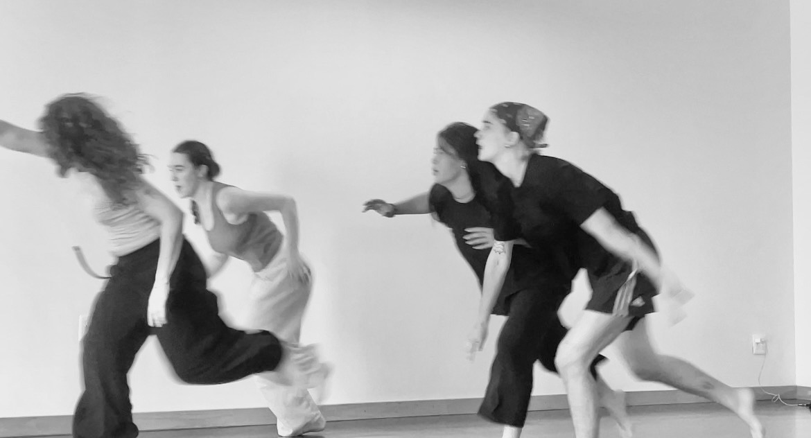 In a black and white photo, four dances are mid-run. The image is slightly blurred, and they are in different moments of stepping forward and reaching.