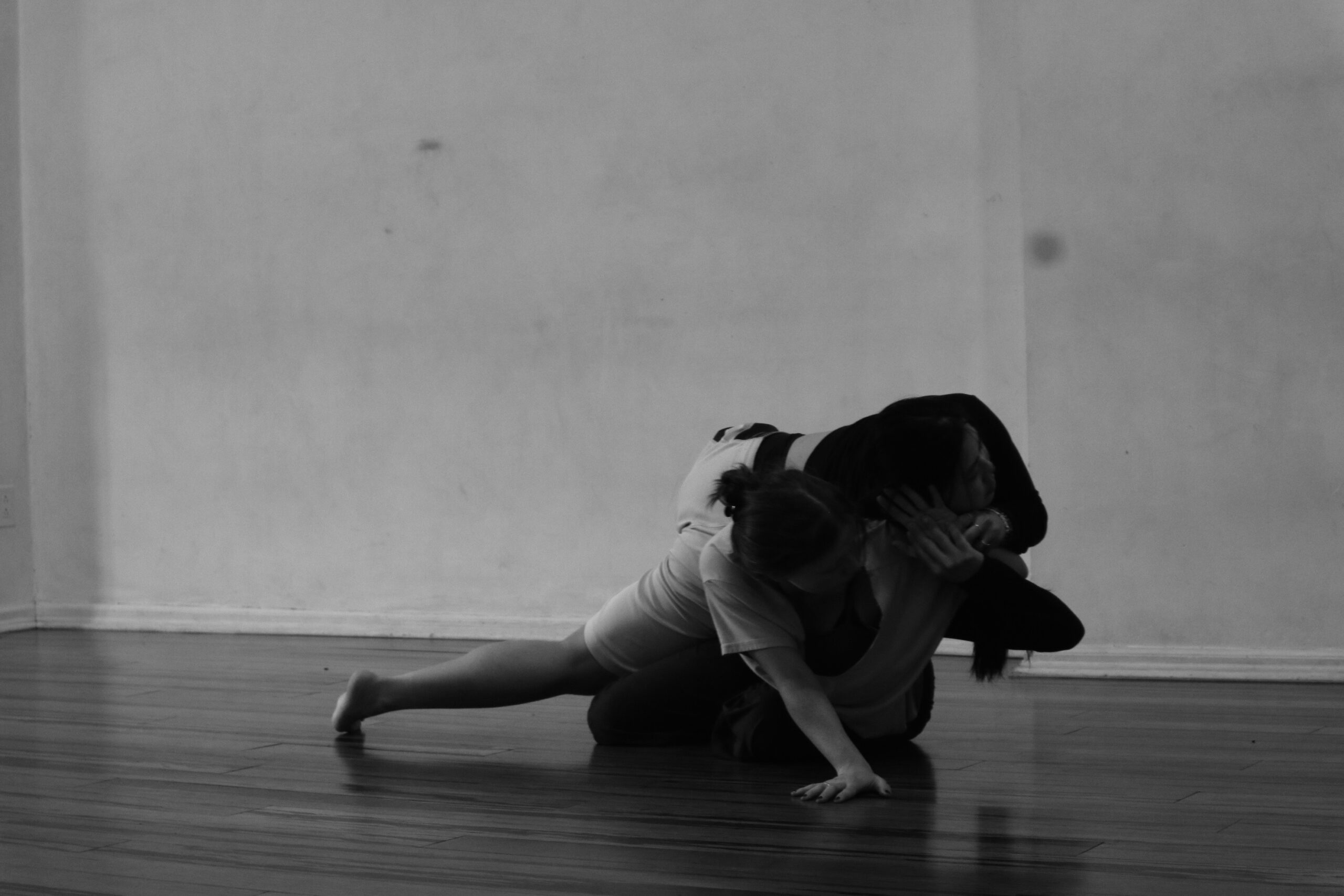 In a black and white photo, two dancers lay one on the ground, one on top of the other. The bottom dancer is supported by their outstretched hand on the ground, while the other touches the dancer's palm on top of them.