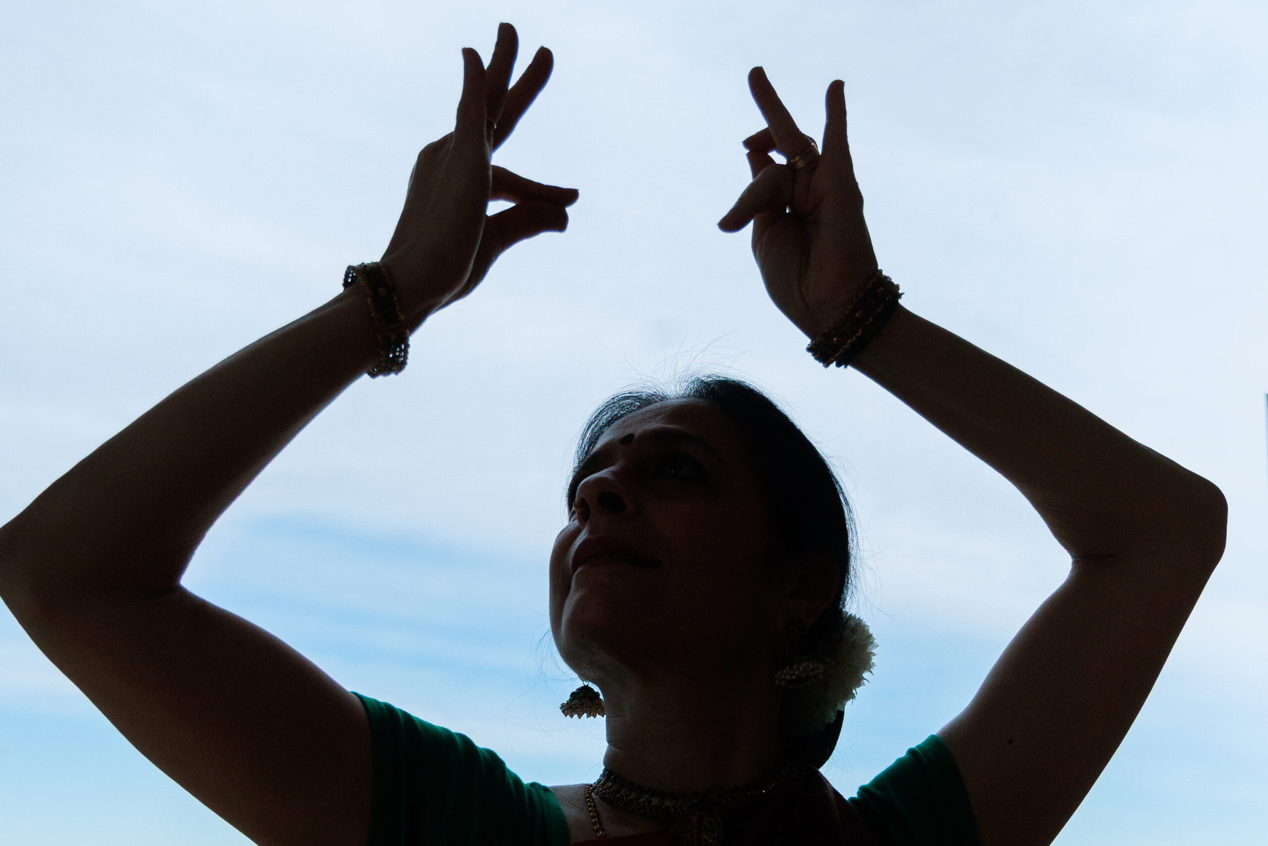 Nalini Prakash is backlit against a blue and white sky, where you can just see her head and arms. She holds two mudra shapes with arms bent, framing her head as she gazes towards her left hand. There is an airy lightness with her crisp gestures that frame a focused gaze.