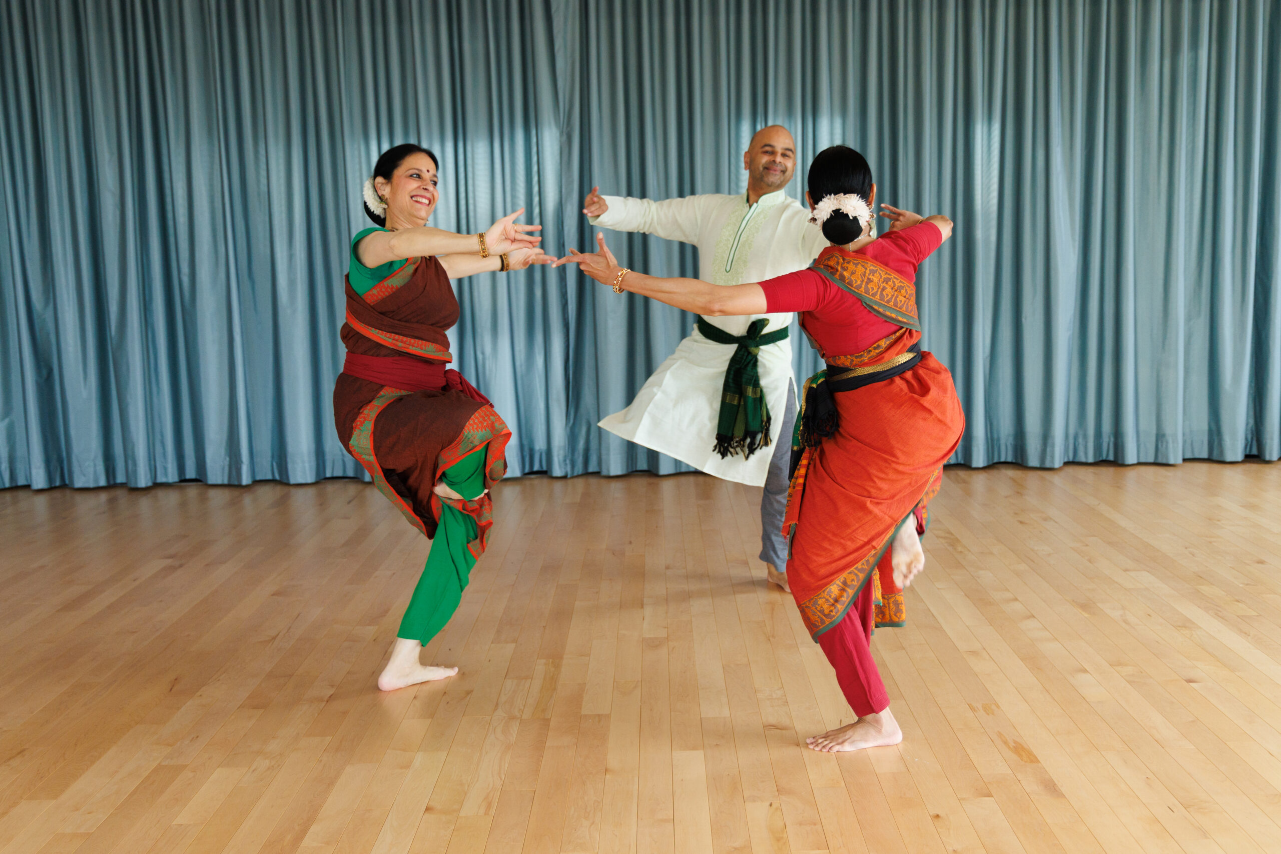 Three dancers, including Nalini Prakash and Vijay Palaparty all face each other in a circle, dressed in vibrant greens, reds and yellows while Vijay wears mostly all white. They are in a dance studio with warm wooden floors. They are all smiling at each other, reaching arms around while balancing on one foot.