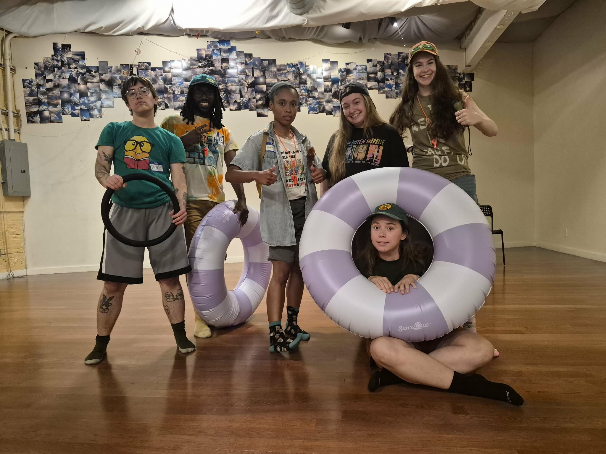 Image Description: Five young would-be campers, mostly smiling, with goofy patterned T-shirts standing in socks on a wood floor displaying supportive thumbs up. One dancer is seated, their face framed by a round lavender and white floaty.