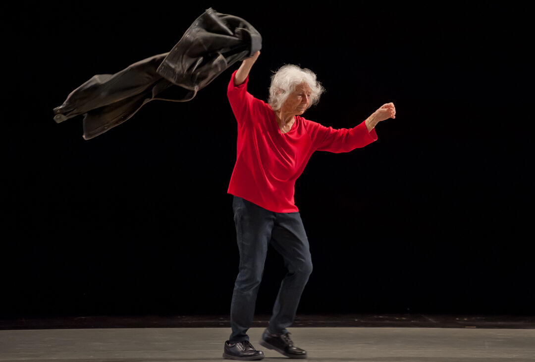 Standing, Simone Forti lifts both of her arms. She wears a foream-length red shirt, and she has short white hair.