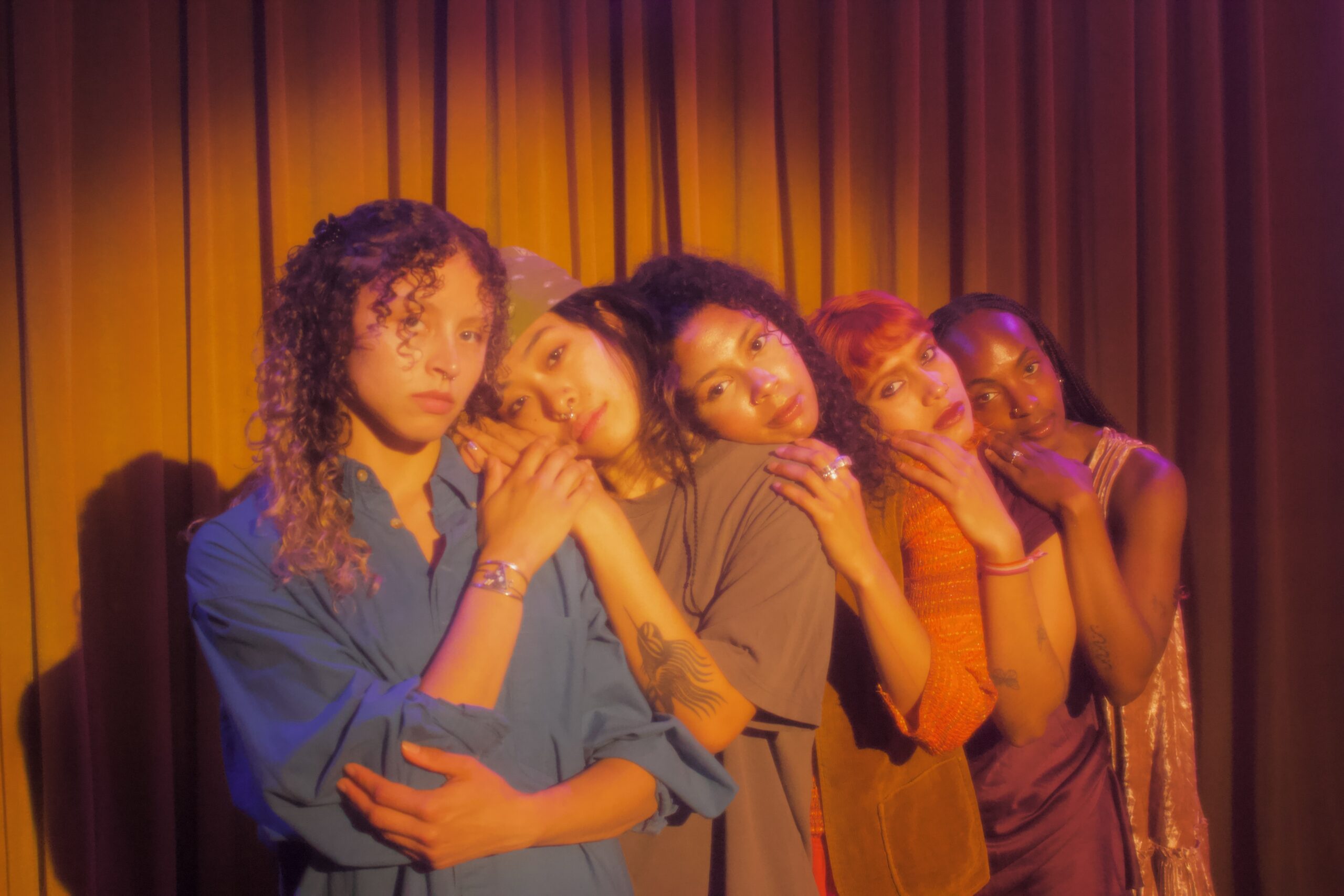 Five dancers rest their cheeks on one another's shoulders in a long, diagonal line. They cup one another's elbows and stare directly into the camera.