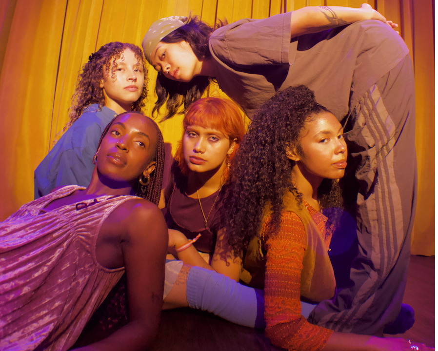 With serious expressions and close proximity to one another, five dancers sit, lean, stoop, and rest casually.