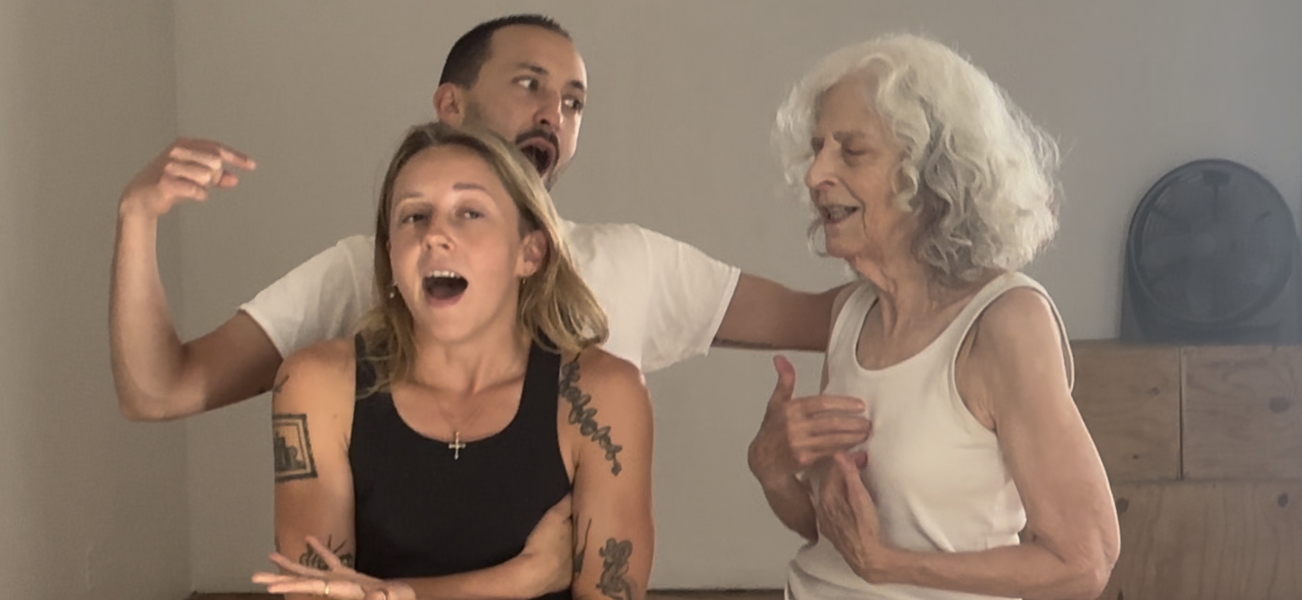 three performers wearing plain white or black tanks/tees are mid gesture as if having a family argument. The performer in the front hunches their shoulders, crosses their arms, and has mouth open, perhaps in the middle of a sentence. Behind them, another performer looks to be yelling and pointing at the third performer, a hand on their shoulder, who has eyes closed, a soft smile, and holds their fingertips to the center of their chest.