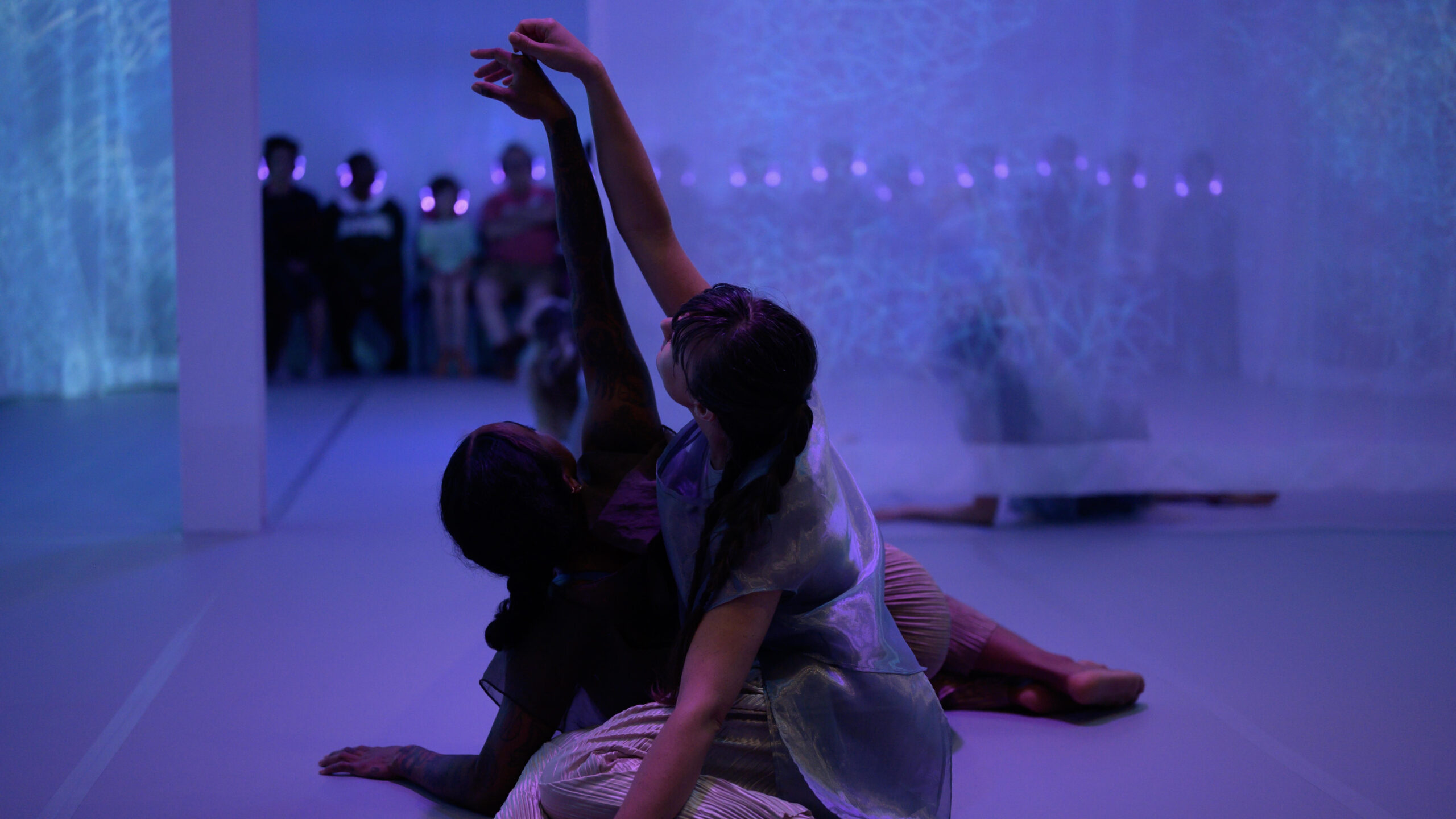 Chloe Marie Newton, a black female dancer wearing a gauzy dark blue tank and Helen Hale, a white female dancer with a long brown ponytail wearing shimmery silver, sit on a light grey marley floor, reclined on one hip with knees gently stacked and one arm intertwined with the other, reaching upwards. They have their backs to us and around and beyond them, we can see pale, cool-toned, sheer fabric. Through and beyond the fabric, we see a row of shadowy-figured audience members, seated and wearing headphones that emit a blue glow.