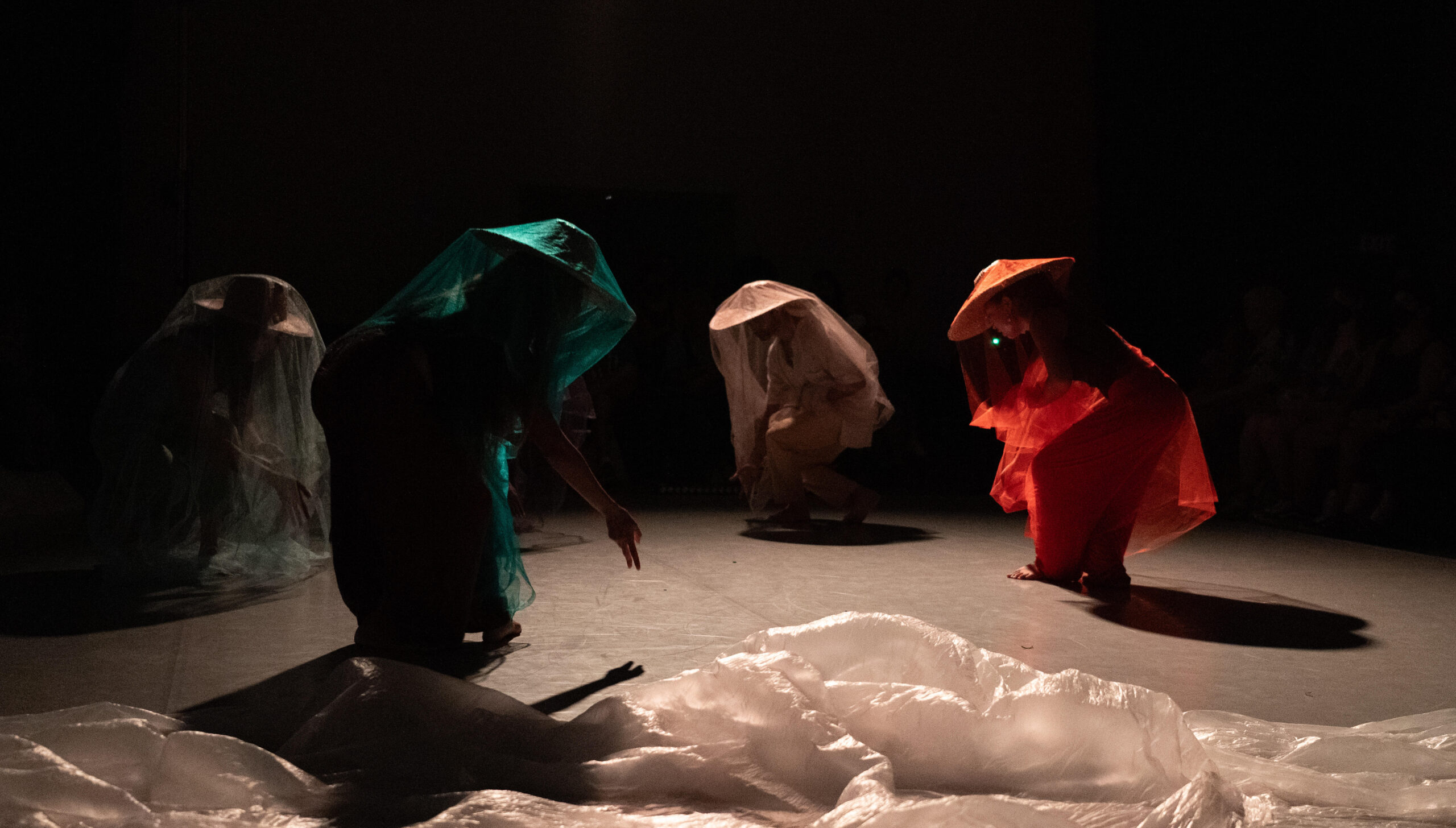 Image Description: Four figures, shrouded in gauzy silver, turquoise, crimson and beige sheer fabric atop their salakots (Asian-Strykers conical hats), crouch forward gesturing to the Earth. Surrounded by blackness, they stand, casting shadow on a light grey marley floor. There is a long piece of white plastic sheeting strewn across the floor in the foreground.