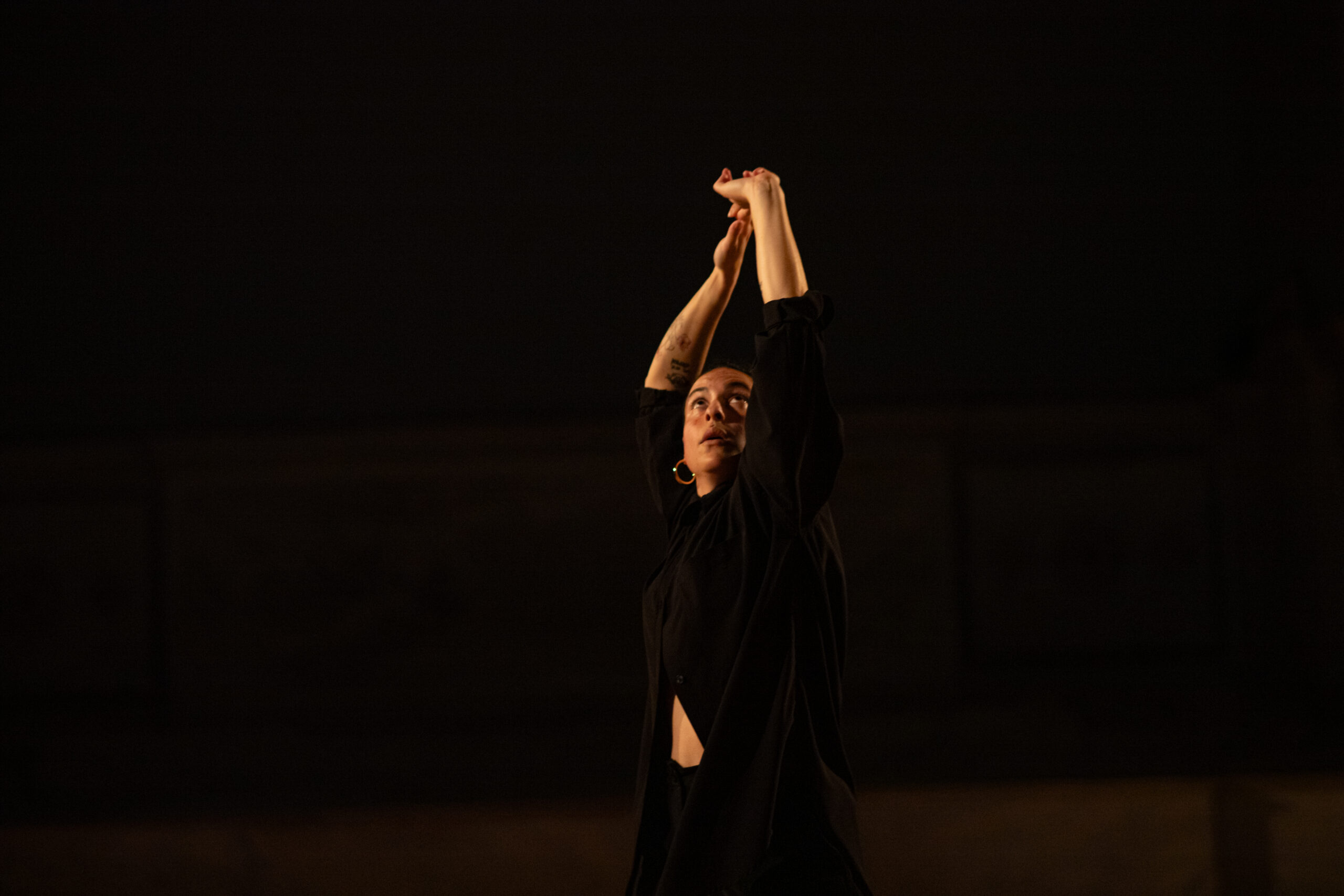 A performance image of Zaquia Salinas, taken during her piece, A Call to Prayer. She wears large hoop earrings and an all-black outfit, with a shirt that wraps and opens around her stomach. Her gaze is directed upwards with a focused expression that manages to feel both wondrous and apprehensive. Her arms snake above her head asymmetrically, almost as if her hands are pushing the sky away.