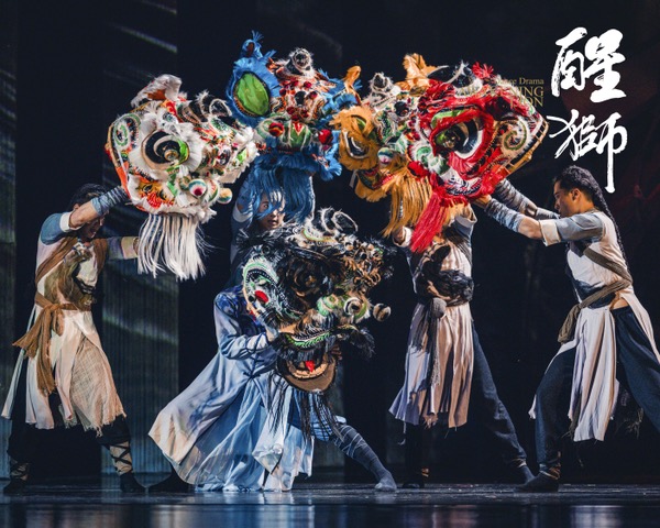 Five dancers in Awakening Lion hold ornate lion heads against a dark stage. White, blue, gold, and red heads form an arc above a black lion head held by one of the story’s heroes (a lion dance champion) in a deep side lunge at the front. The performers wear layered, earth-toned tunics with wrapped forearms and lower legs.