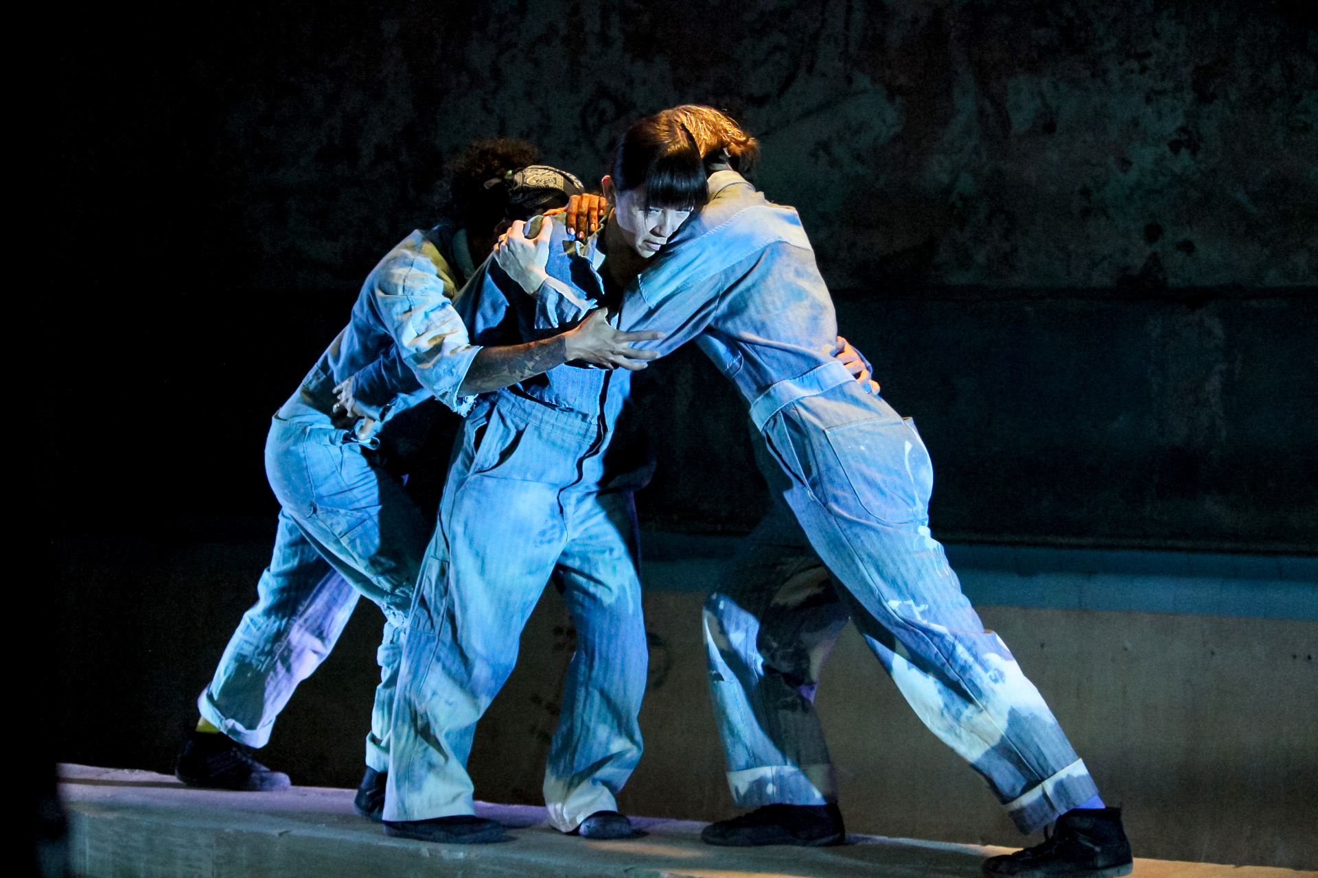 Three women in faded blue and white jumpsuits stand supporting or leaning into each other. Marcie Mamura stands in the middle; she looks down and away, the only face visible in the group. Chloe Marie embraces Mamura from behind. Katherine Kiefer Stark supports Mamura from the front, also in an embrace. The three women create a quiet, intimate, peaceful tableau. Bright light illuminates their bodies against a dark, shadowed background.